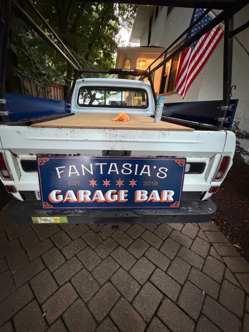 The back of a white pickup truck parked on a brick driveway, with a sign that reads "FANTASIA'S GARAGE BAR EST. 2019." The truck bed has a large wooden surface, a can of spray paint, and an orange cloth. An American flag hangs from the house nearby.