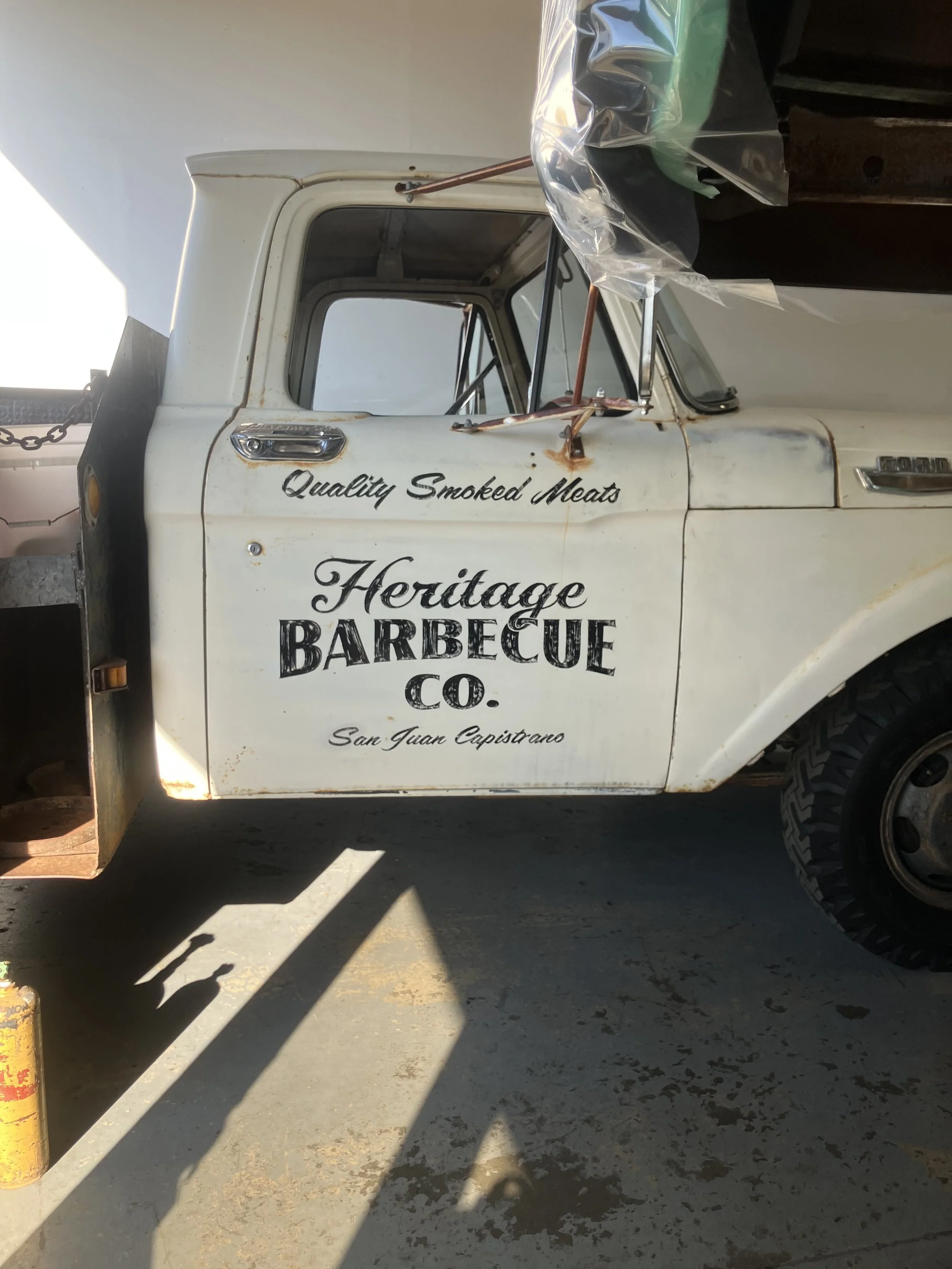 Part of a vintage truck with a white body and black lettering advertising Heritage Barbecue Co. in San Juan Capistrano. The truck has some rust spots, a side mirror, and a large black tire visible. The ground is concrete with shadows cast by yellow b