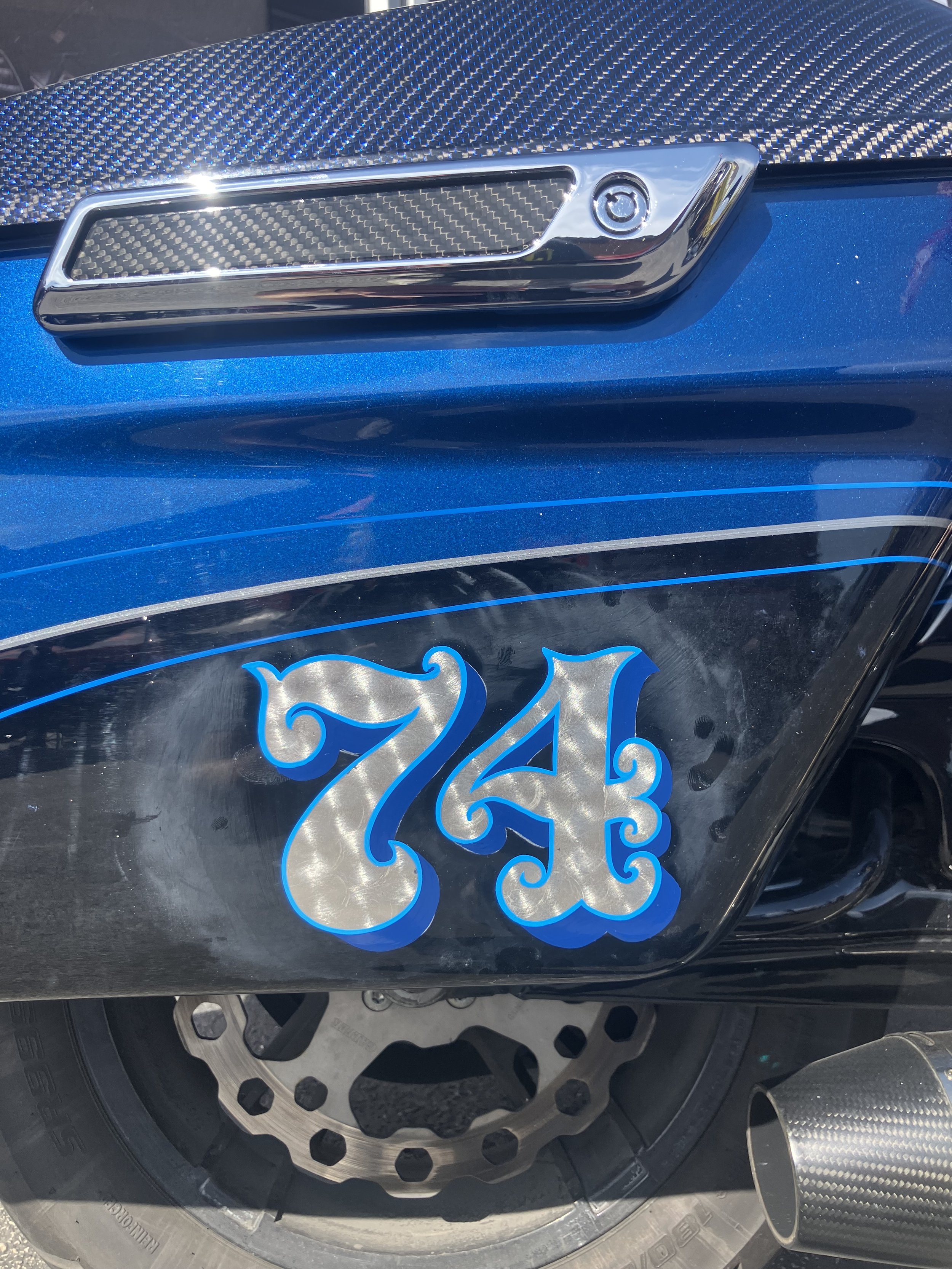 Close-up of a motorcycle front wheel with a blue metallic body and custom decorative design, including carbon fiber accents and a stylized blue and silver logo.