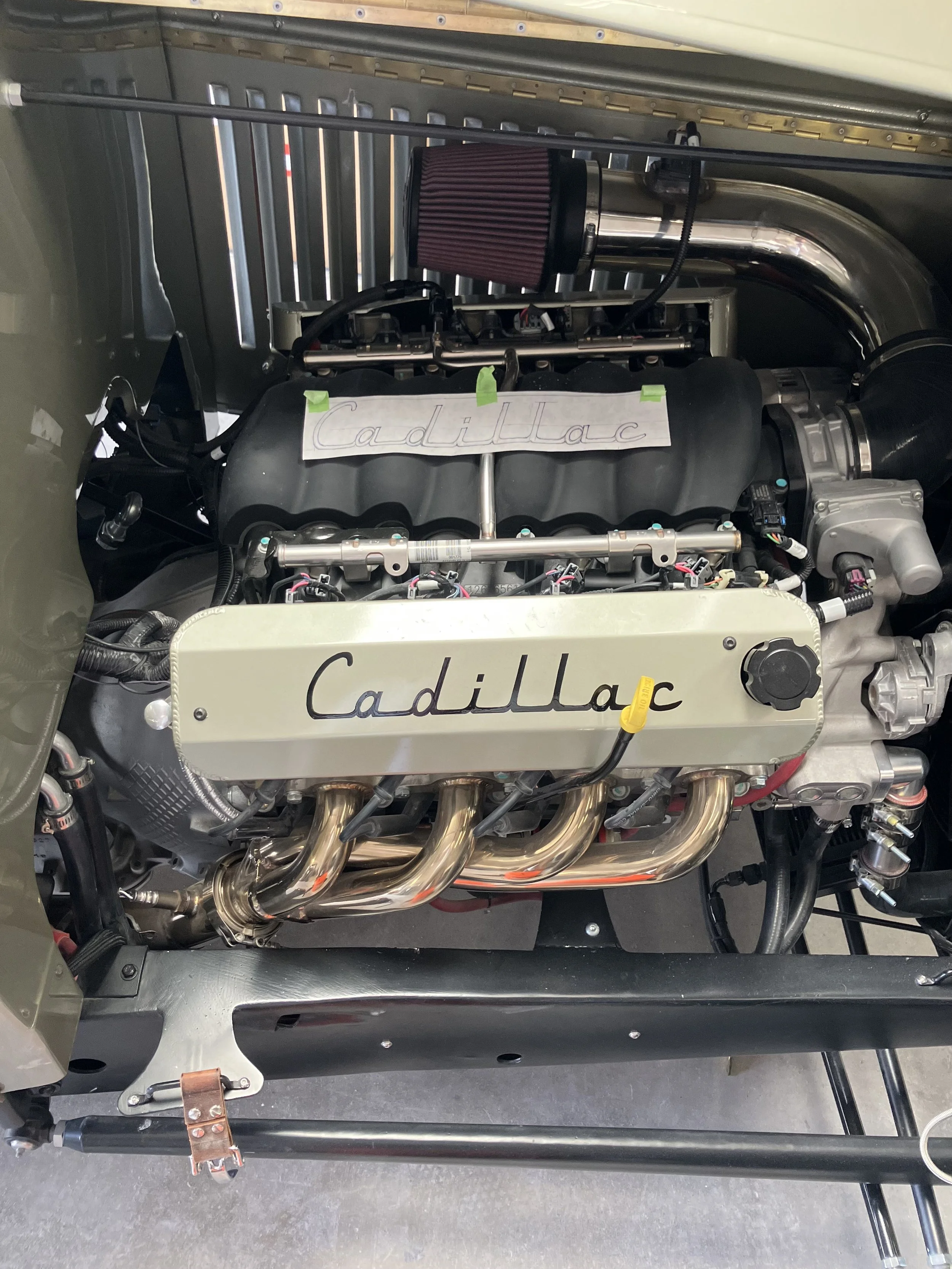 Close-up view of a car engine bay with a labeled valve cover that says 'Cadillac,' a red air filter, and shiny exhaust headers.