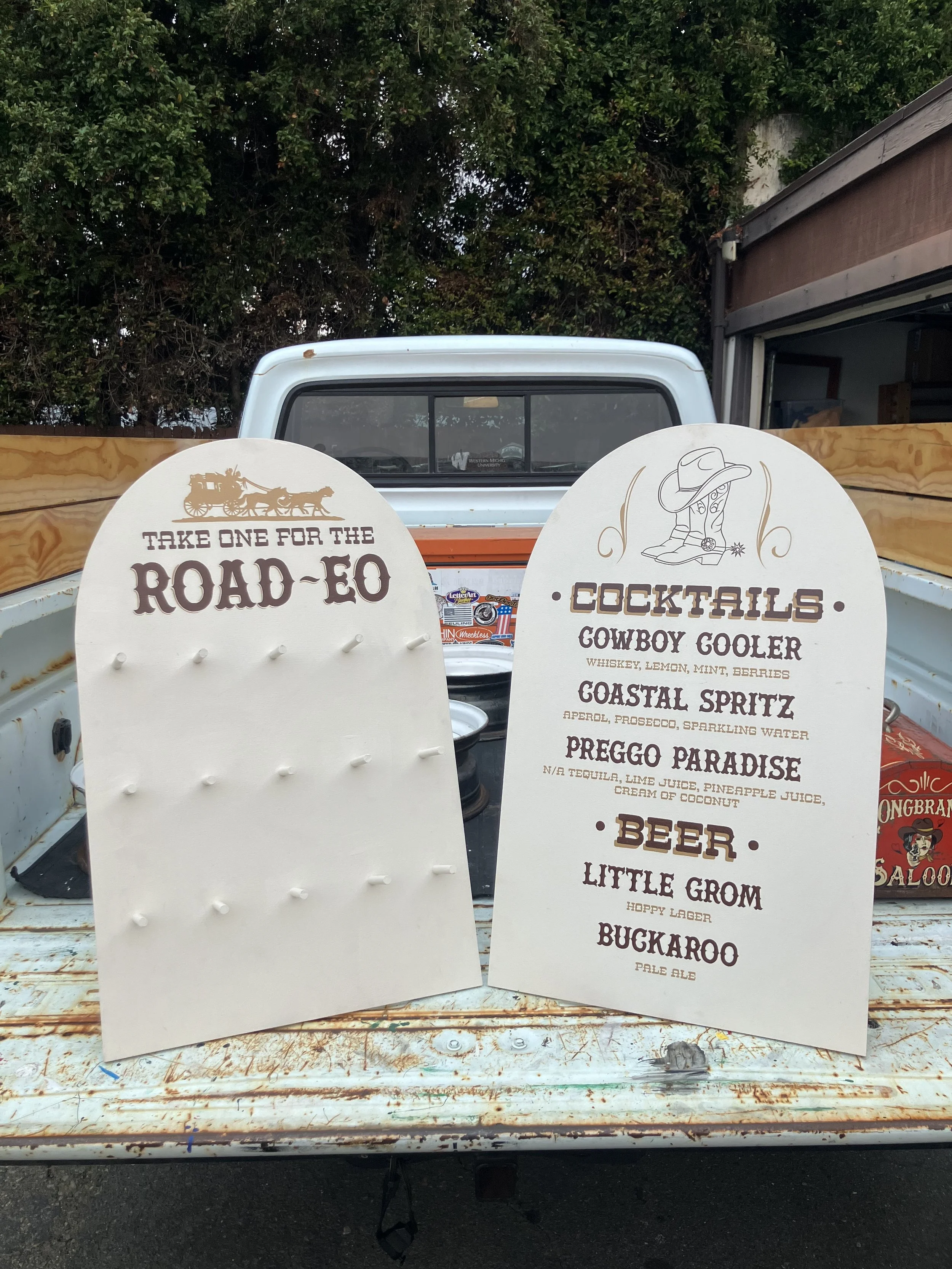 Two white menu boards with brown text and illustrations, listing cocktails such as Cowboy Cooler, Coastal Spritz, and Preggo Paradise, placed in the back of a pickup truck with a wooden fence and trees behind it.