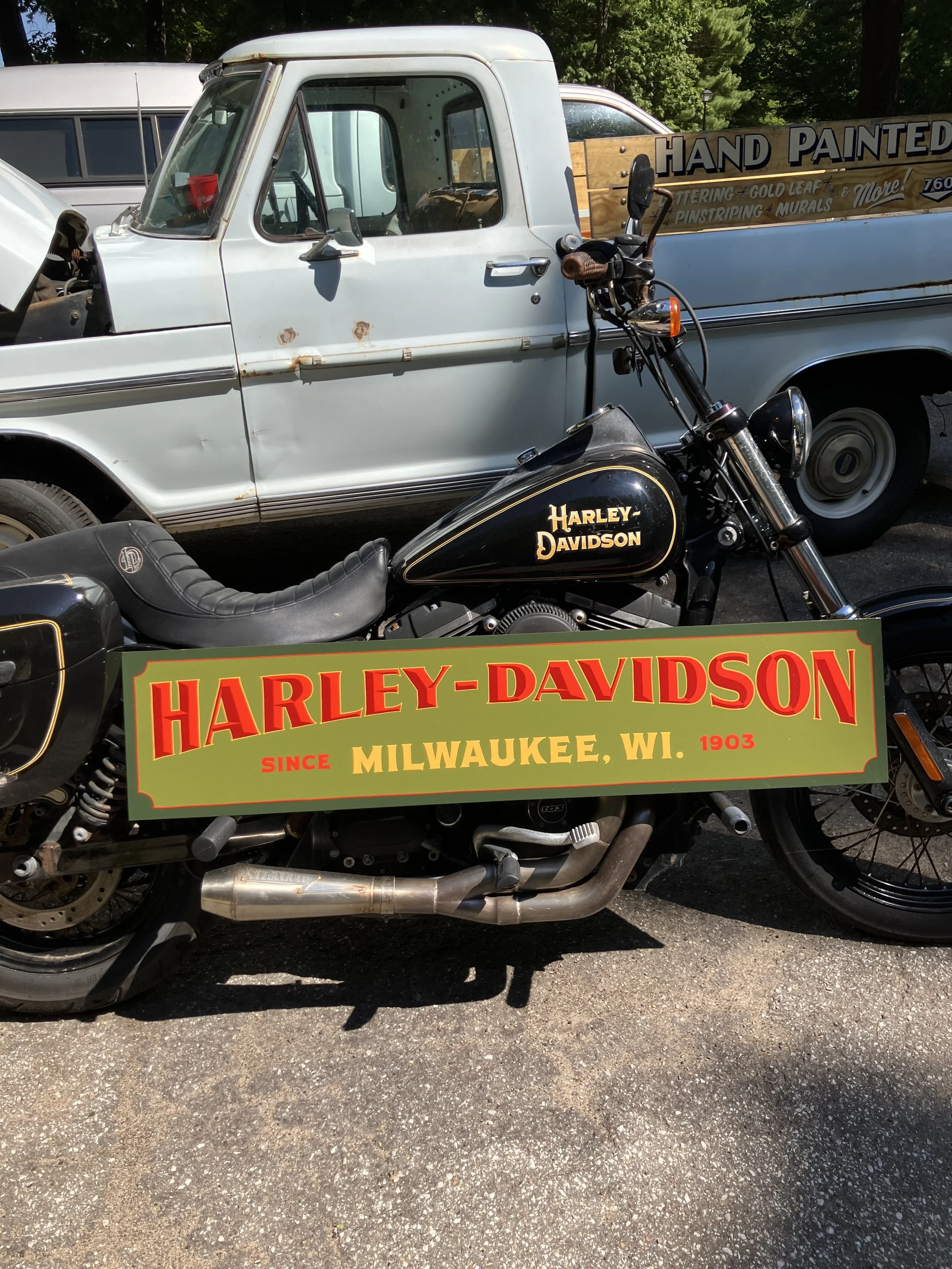 A vintage Harley-Davidson motorcycle with a black fuel tank and gold lettering, parked in front of an old, white pickup truck with a wooden sign in the bed reading, "Hand Painted" and additional text about painting services, in an outdoor setting wit