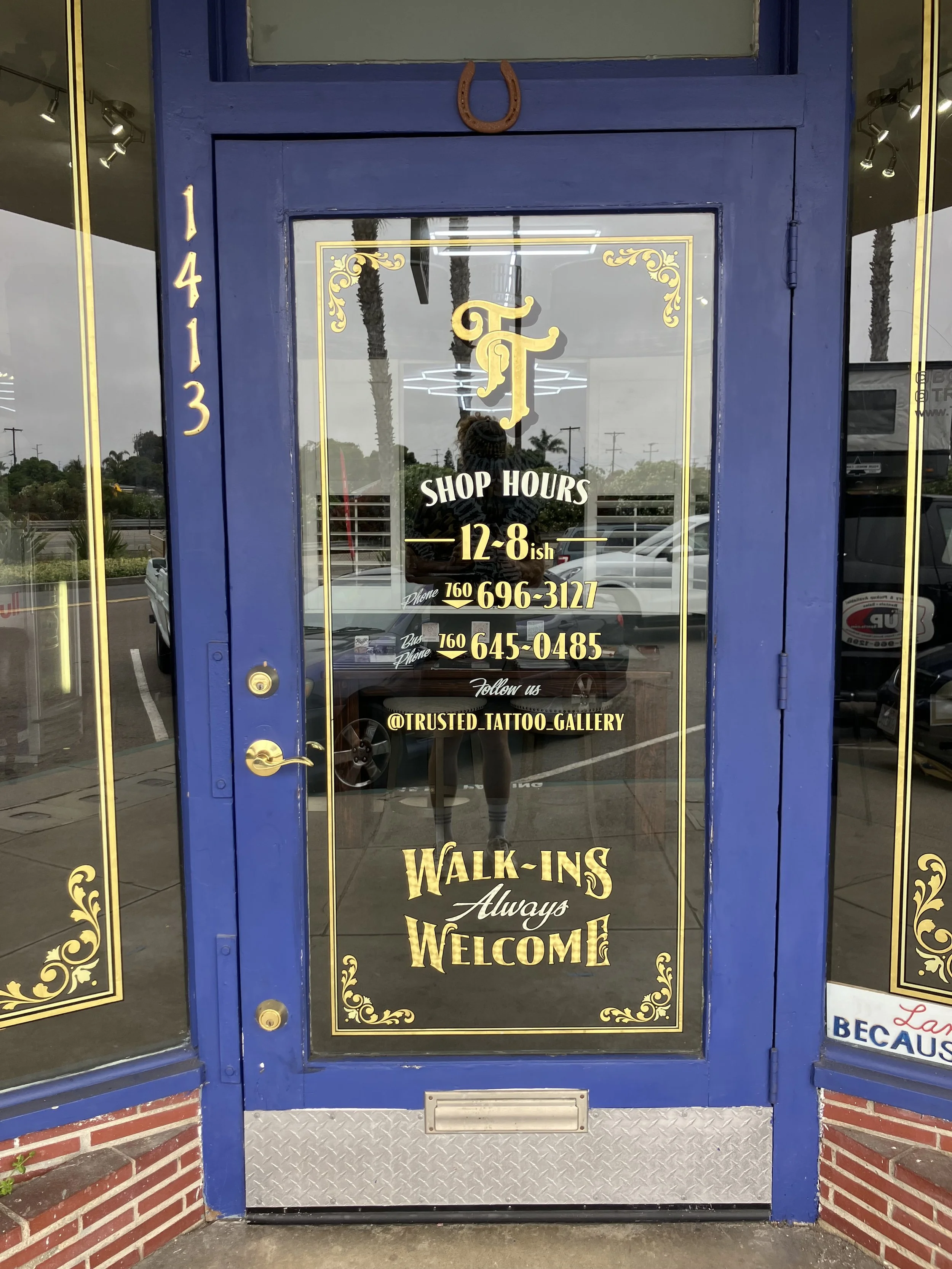 Blue door with gold accents and text advertising tattoo shop hours, contact information, and welcoming message, reflecting in the glass.