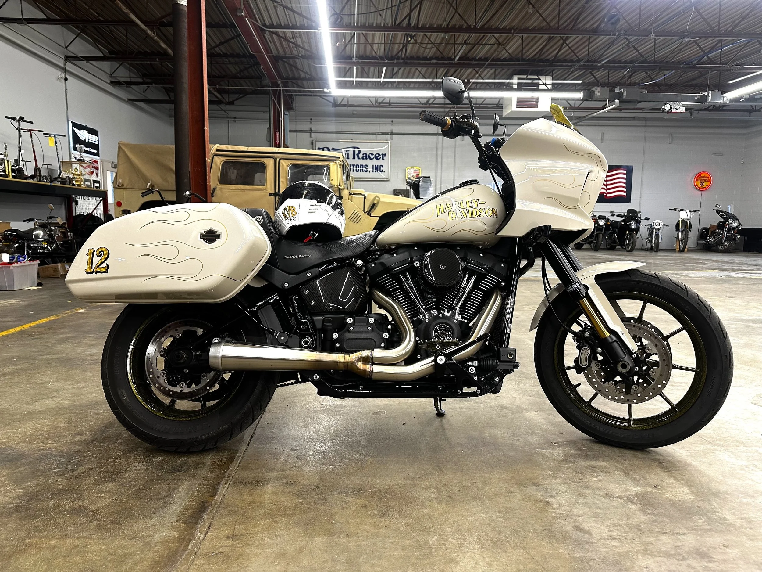 A white Harley-Davidson motorcycle with side saddlebags displaying the number 12 and flames, parked inside a warehouse or showroom with other motorcycles and vintage items in the background.