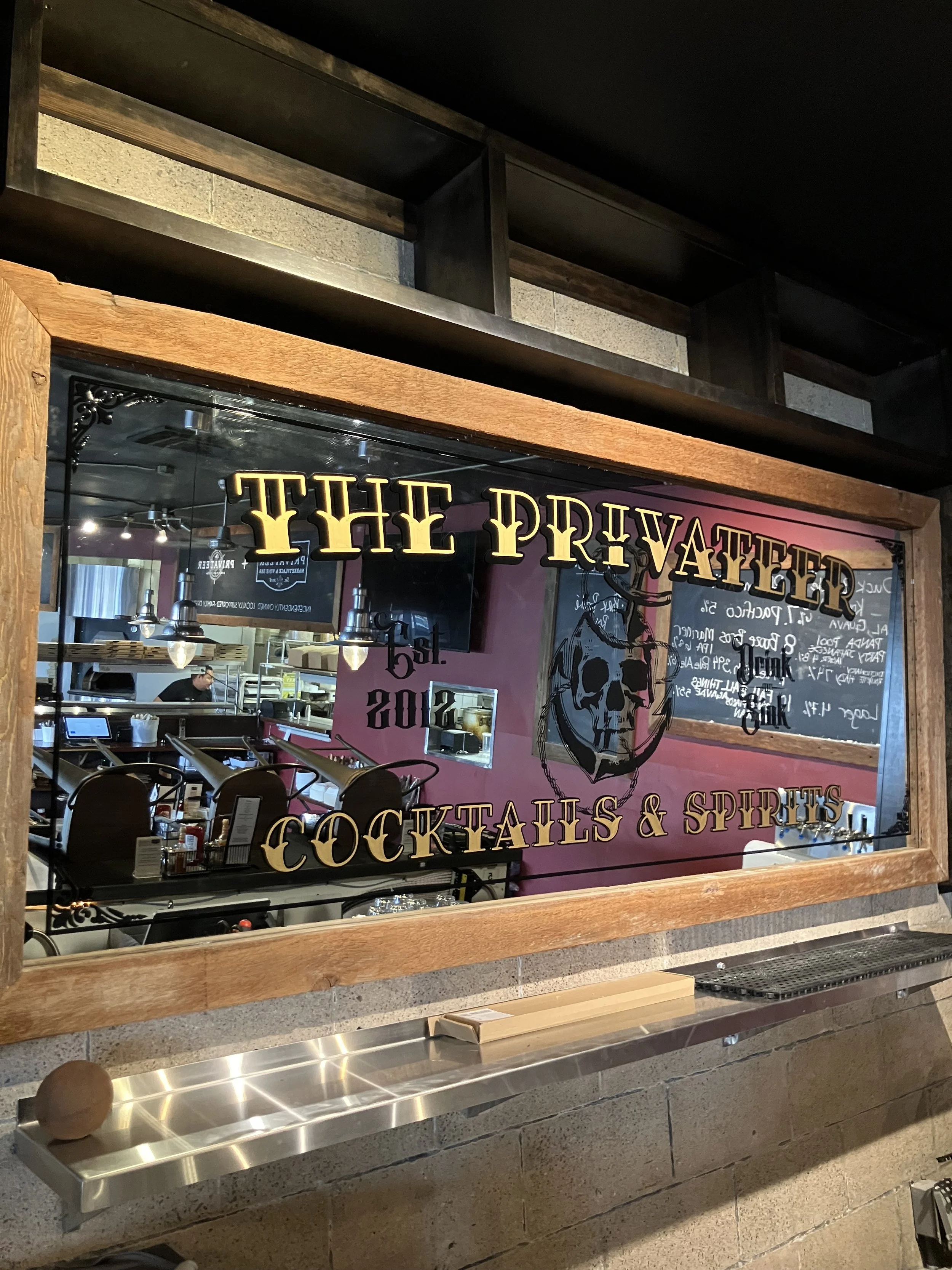 A mirror with the words "The Private" and "Cocktails & Spirits" written on it, reflecting the interior of a restaurant or bar with a menu board and decorations, and a chef working in the kitchen.