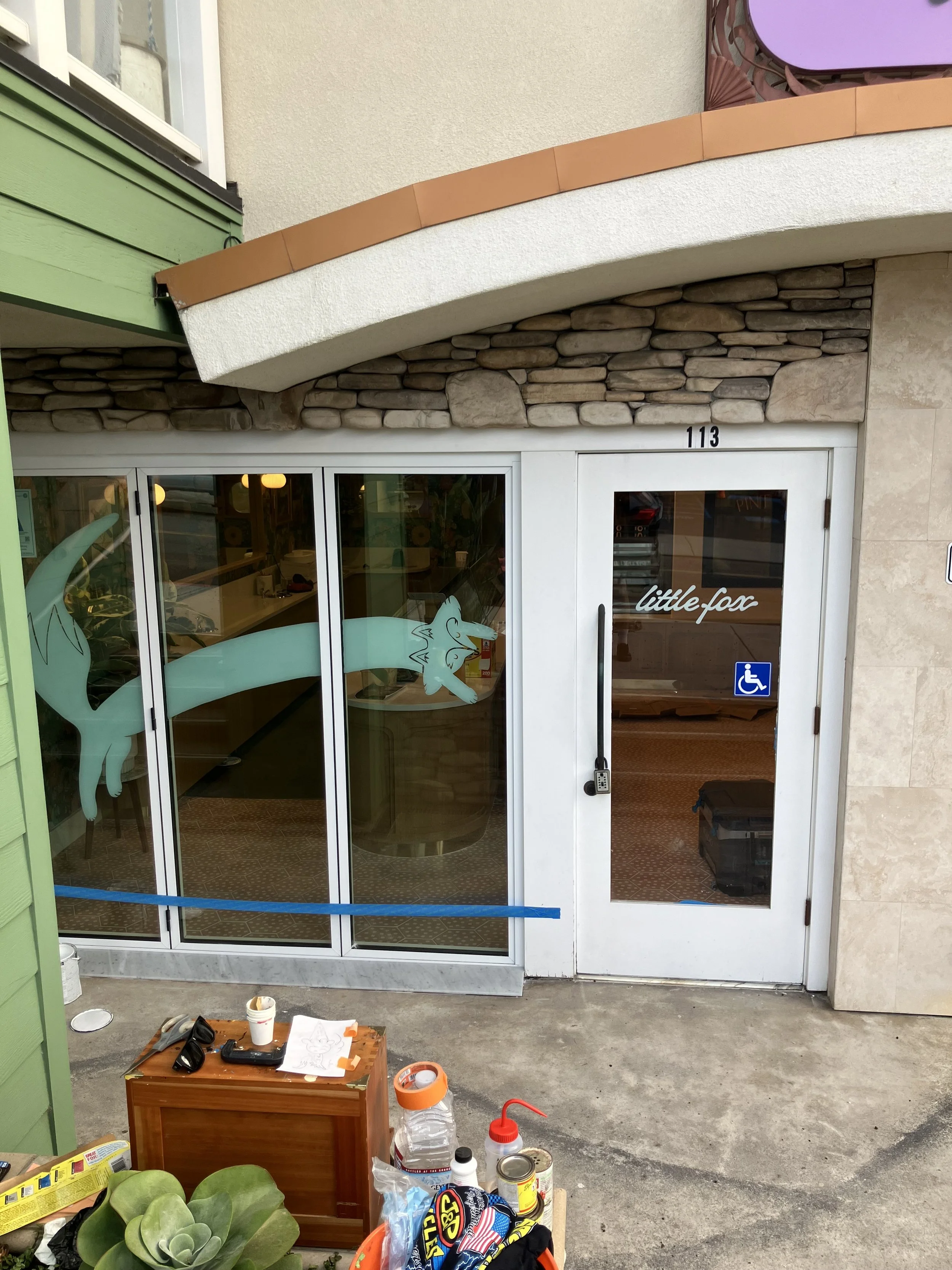 Entrance to a shop with glass doors featuring a fox logo and a handicap sign, surrounded by various clutter including bottles, cans, and plants.
