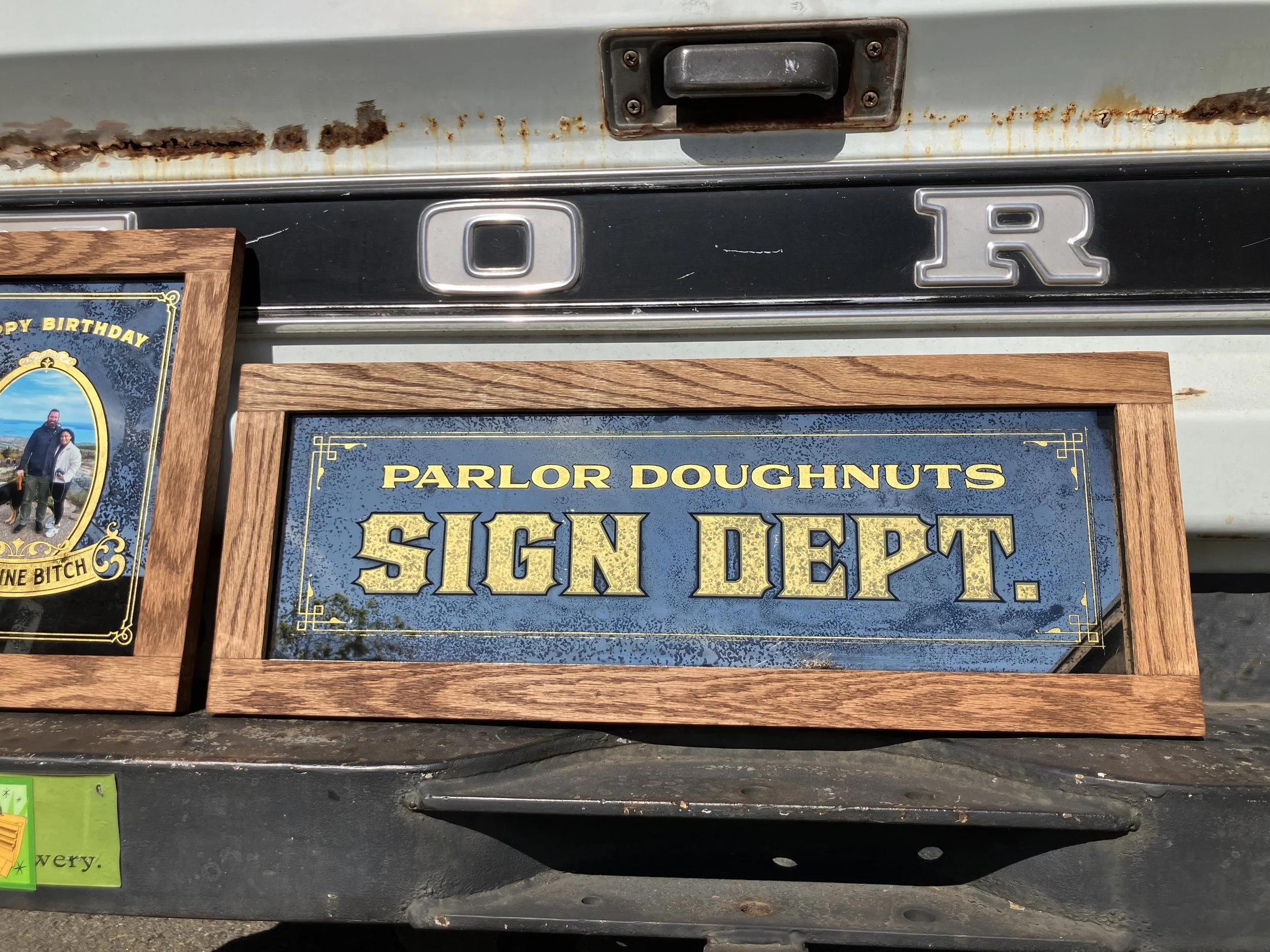 Close-up of the back of a vehicle showing a rusted metal surface, a black handle, and a vintage license plate that reads 'SIGN DROP' framed in wood, with part of the phrase 'PARLOR DONUTS' visible.