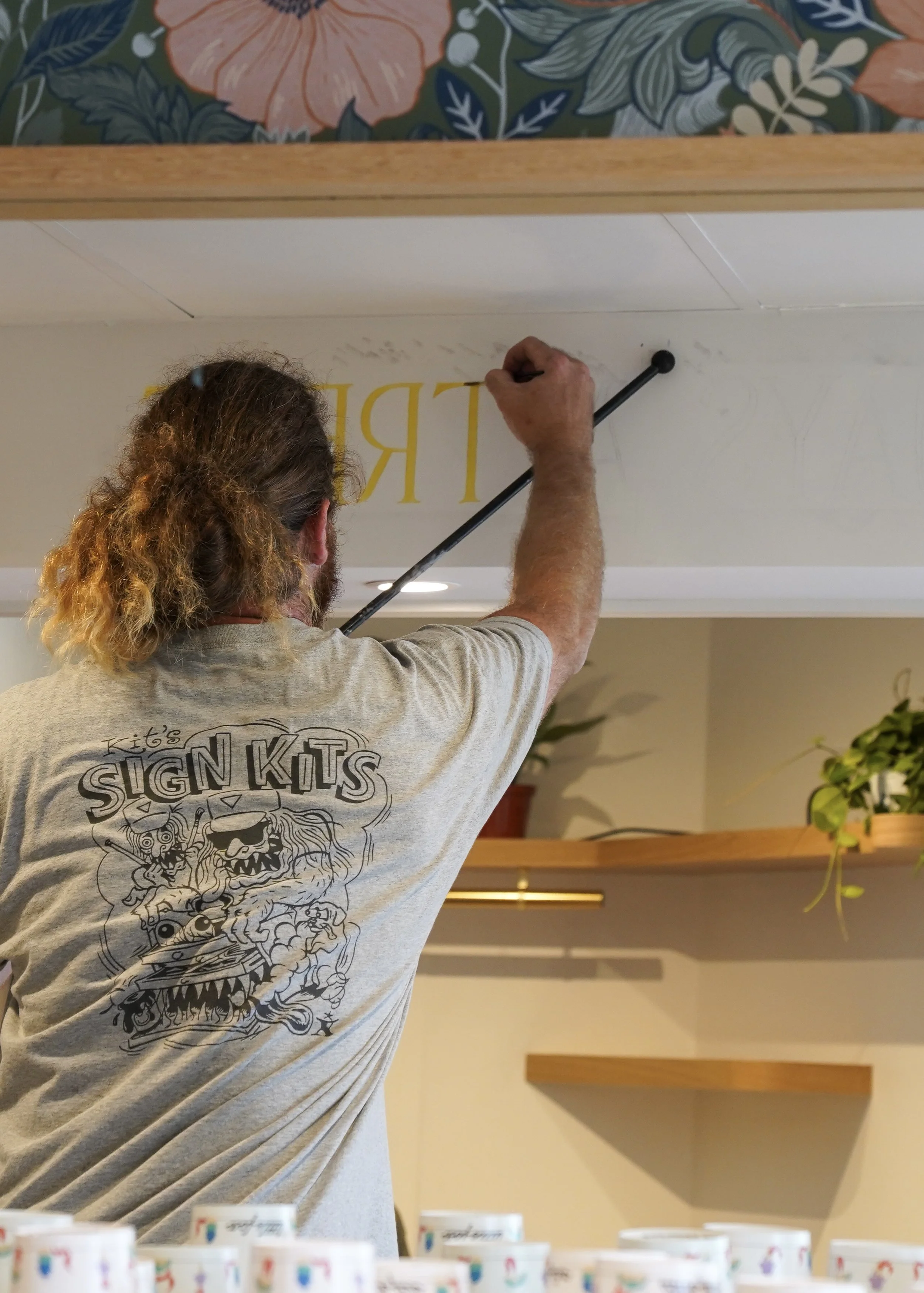 A man with long, curly hair viewed from behind, wearing a gray T-shirt with a graphic design, is chaining a yellow letter onto a wall. The wall features partially visible large yellow text, and in the foreground, there are several colorful cups lined up.