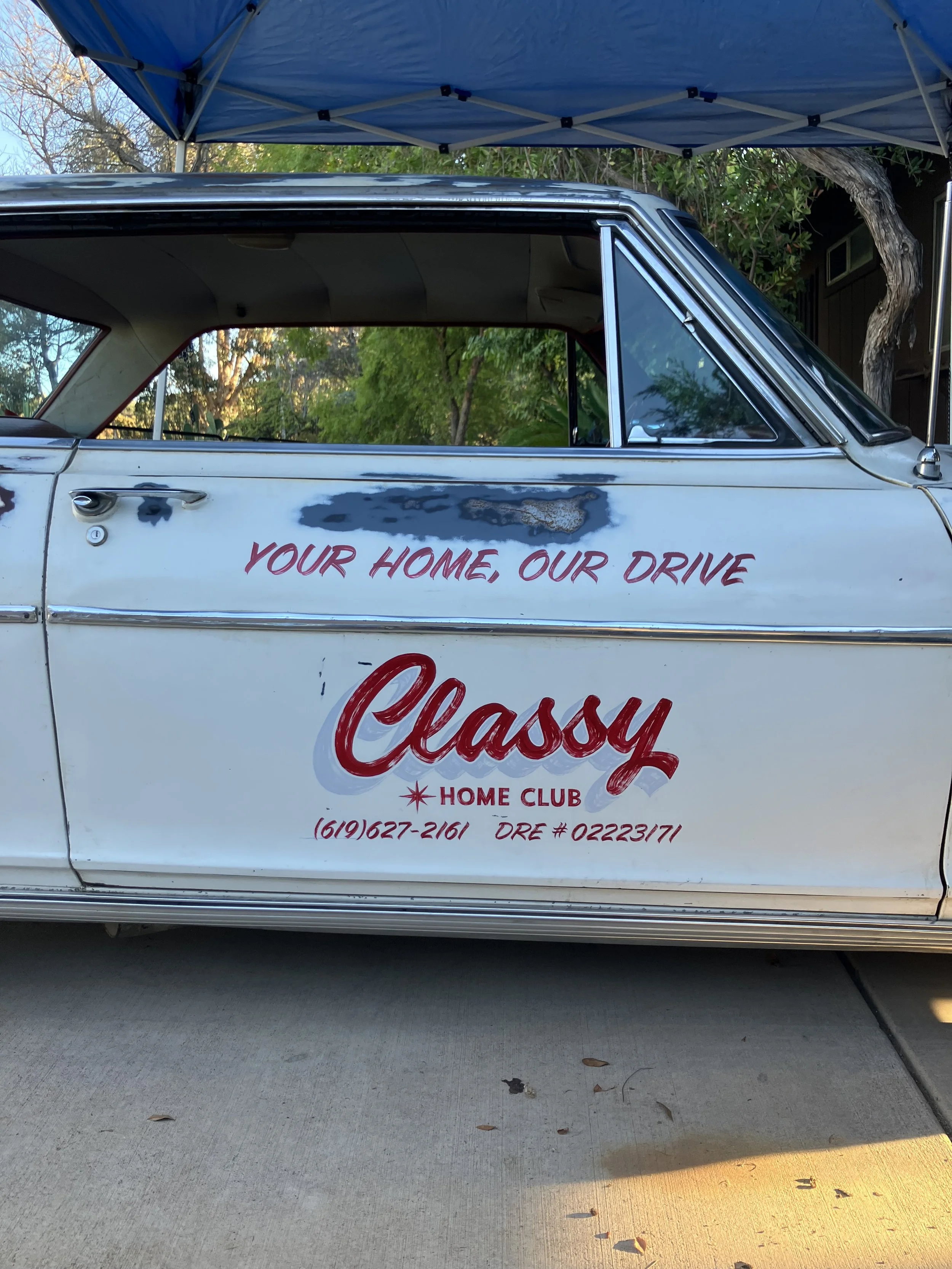 Side view of a vintage white car with the words "Your Home, Our Drive" and "Classy Home Club" written on the door, parked under a blue canopy with trees and a building in the background.
