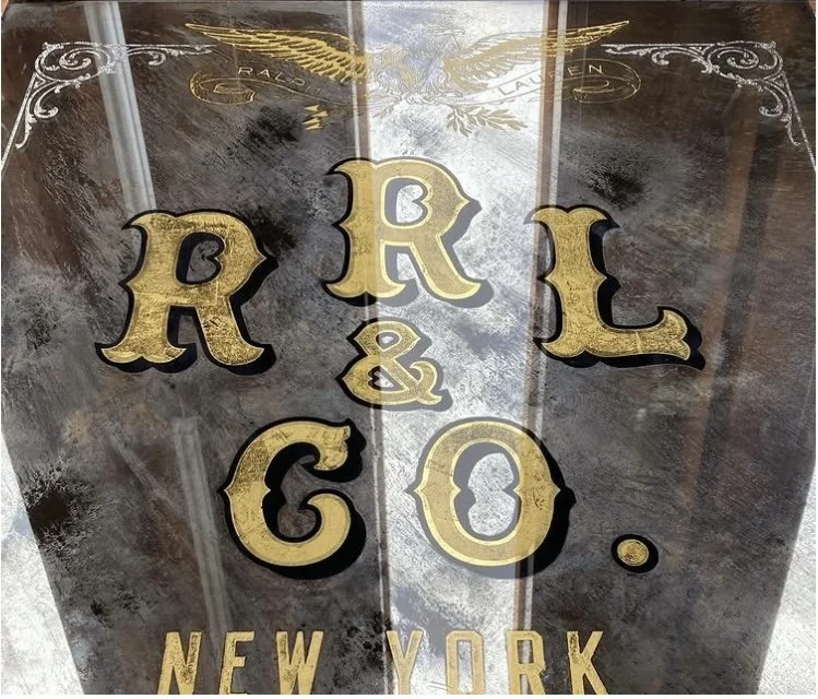 A decorative sign with the word 'RACK & CO.' in large gold letters, and 'NEW YORK' in smaller gold letters below, set against a rustic wooden background.
