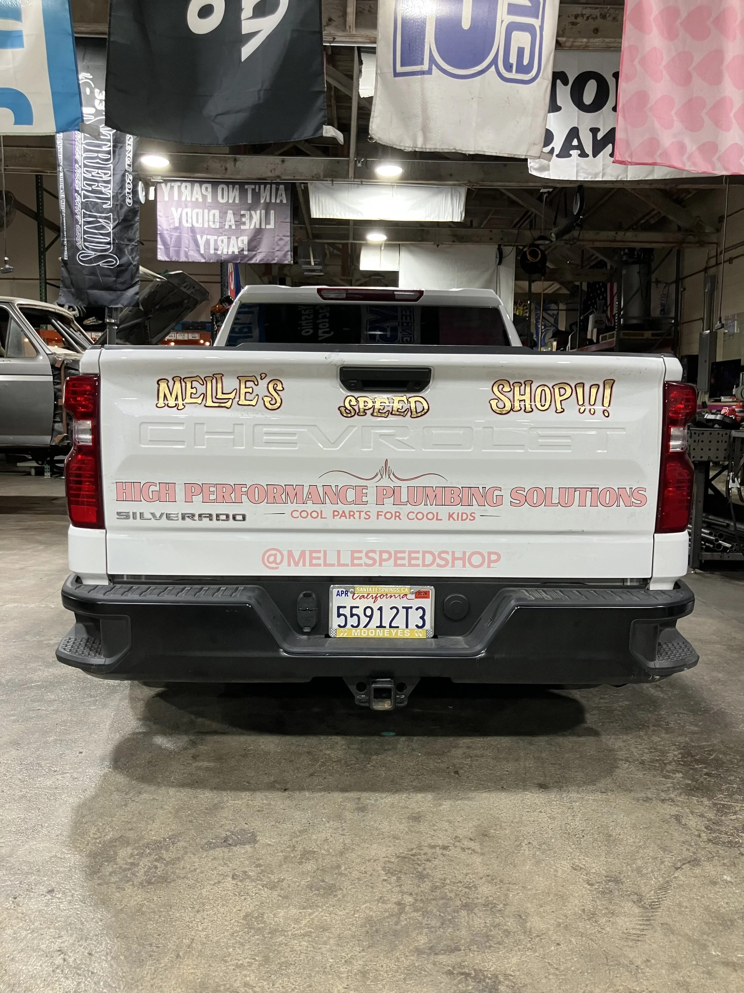 The back of a white Chevrolet Silverado truck with signage for Melle's Speed Shop, a high-performance plumbing solutions business. The truck is parked in an indoor garage with banners hanging from the ceiling.