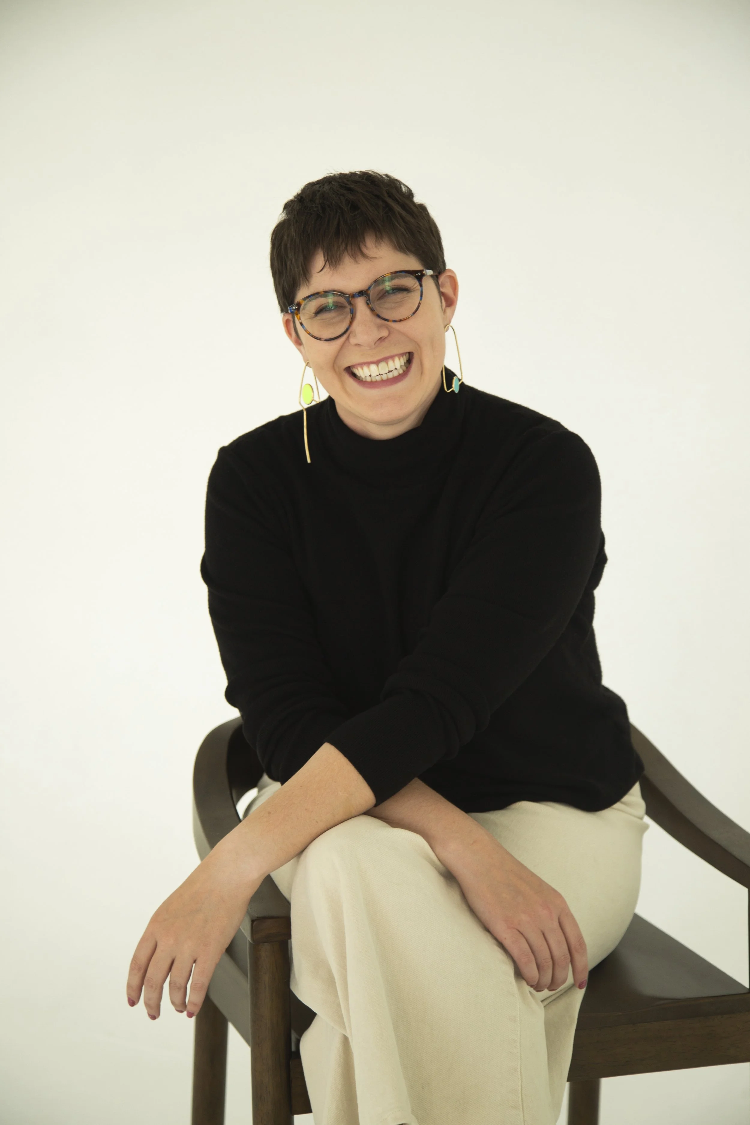A woman with short dark hair, glasses, and hoop earrings smiling while sitting on a wooden chair against a plain white background.