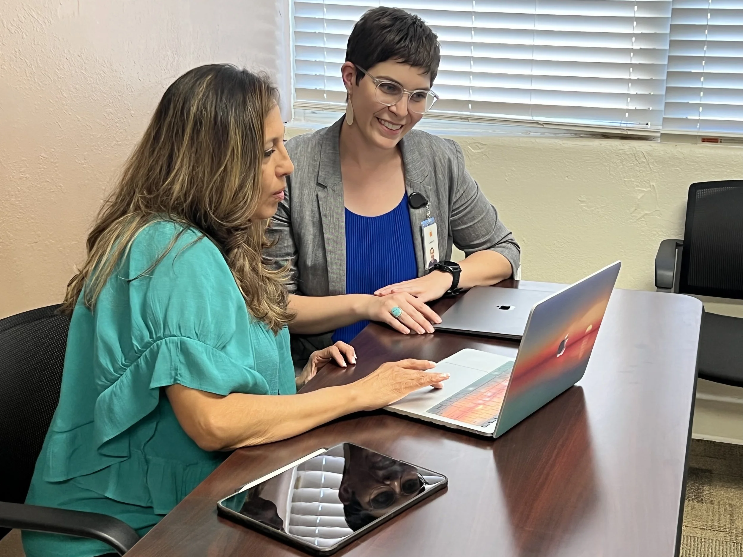 Two women sitting at a desk with laptops, having a discussion in an office with blinds covering the windows.