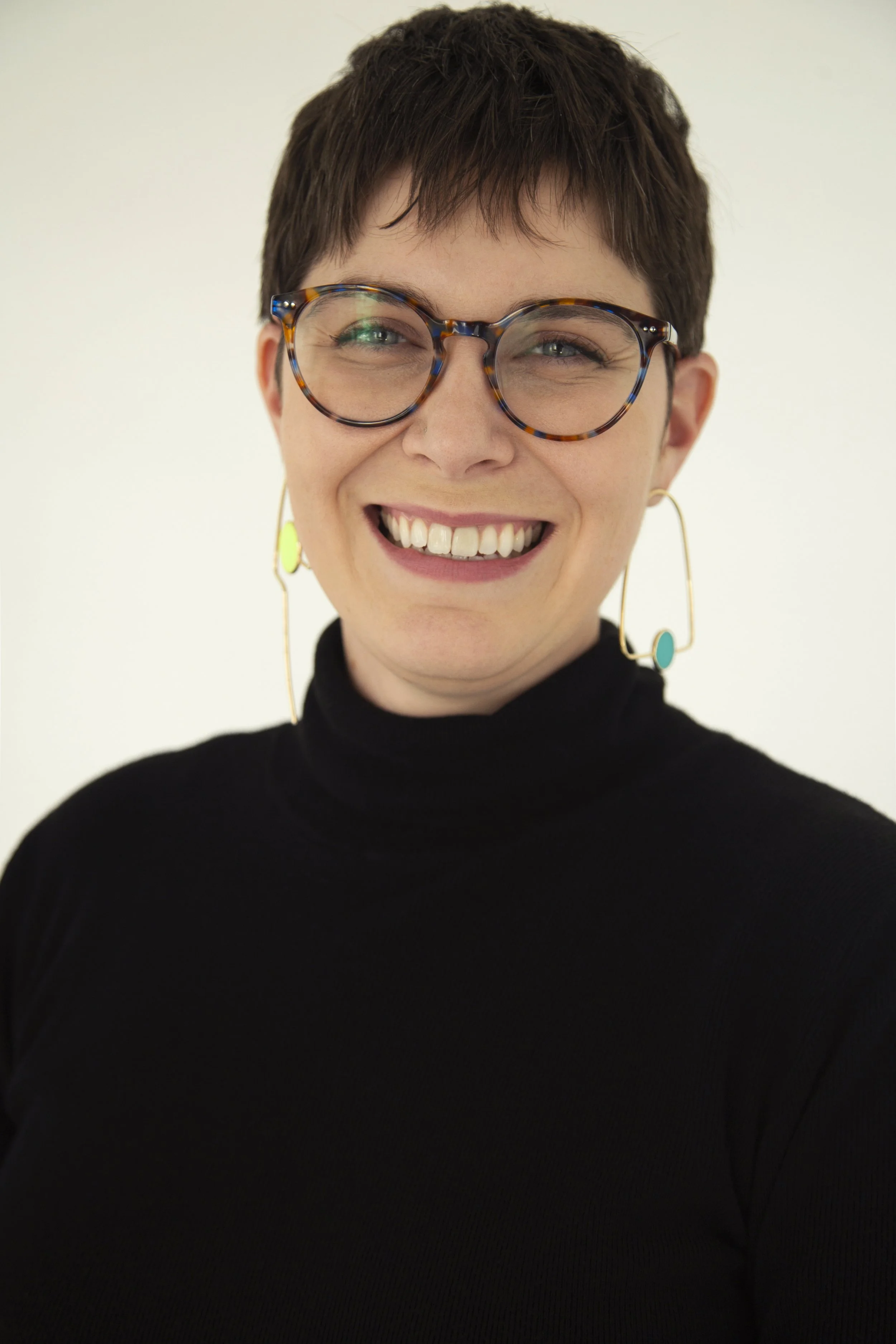 A woman with short dark hair, wearing tortoiseshell glasses, large colorful earrings, and a black turtleneck, smiling at the camera against a light background.