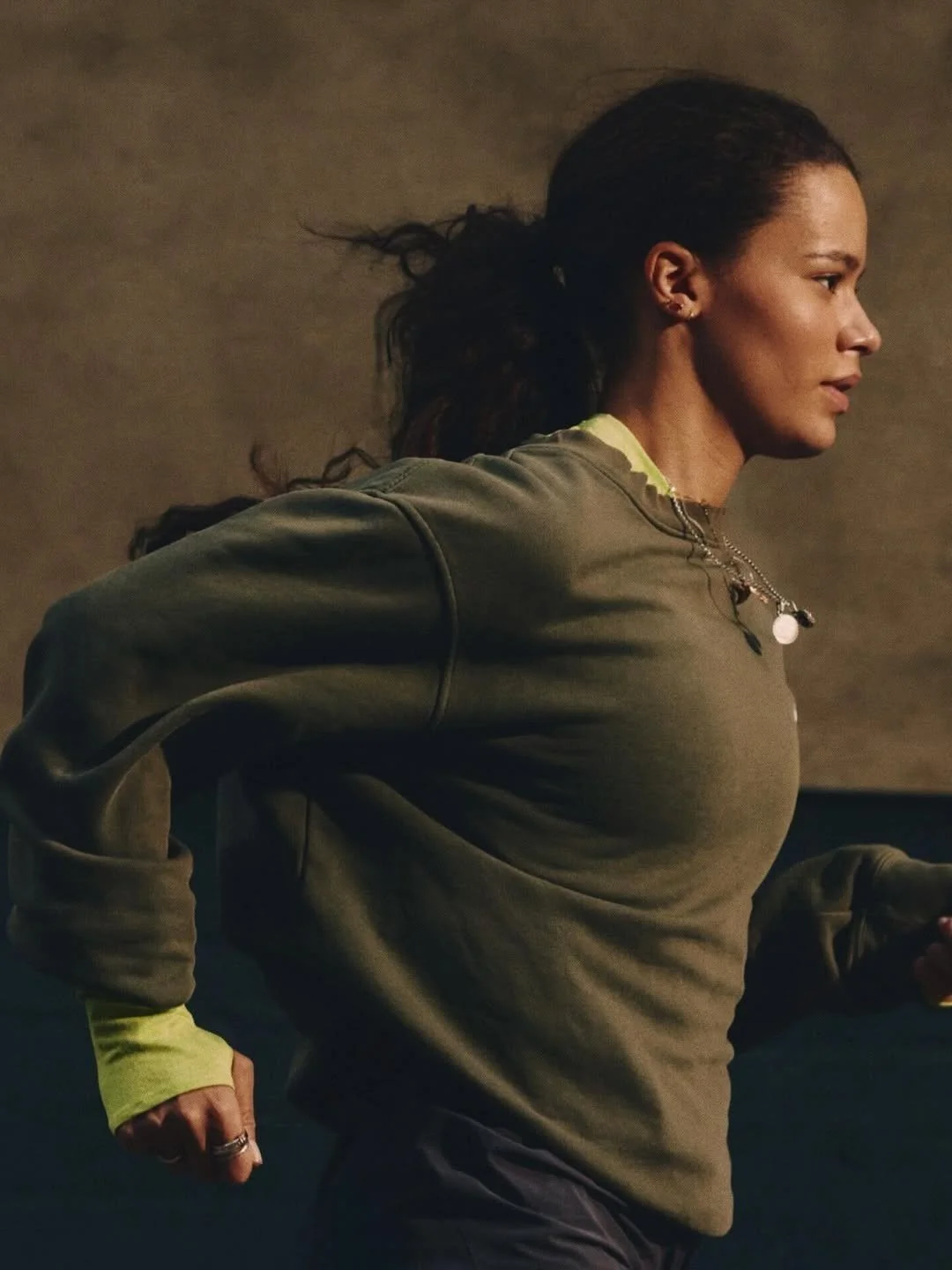 A woman with long dark hair pulled back, wearing an olive green sweatshirt and yellow athletic sleeves, running indoors.