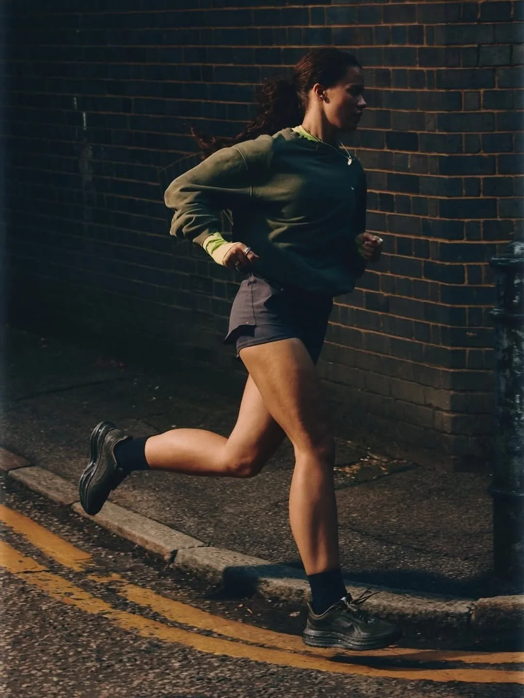 A woman running along a dimly lit street at night, wearing a green jacket, black shorts, black socks, and running shoes.