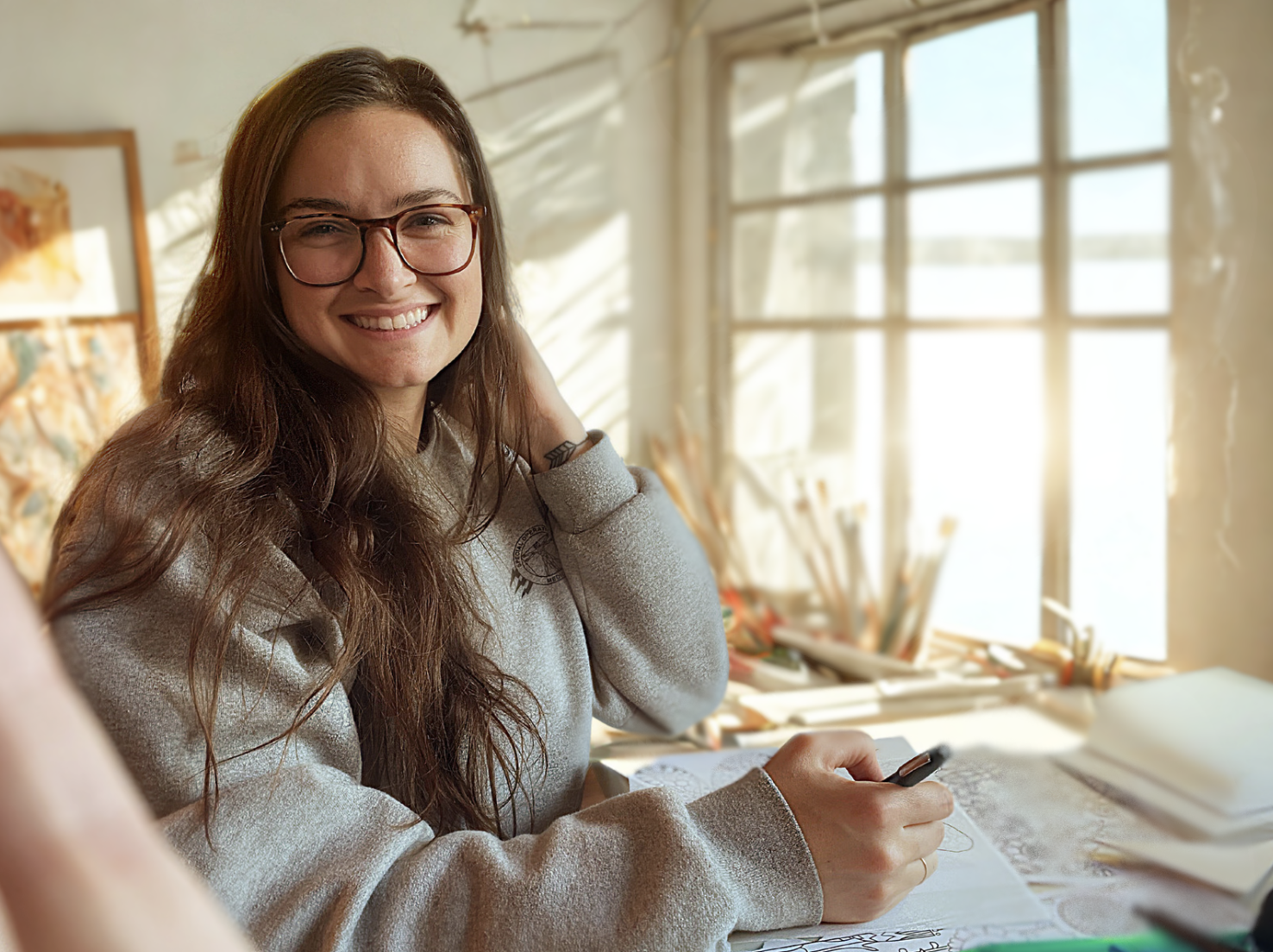 A young woman with long brown hair, glasses, and a light gray sweatshirt smiling at the camera while sitting at a desk filled with papers, sketchbooks, and art supplies, in front of a large window letting in sunlight.