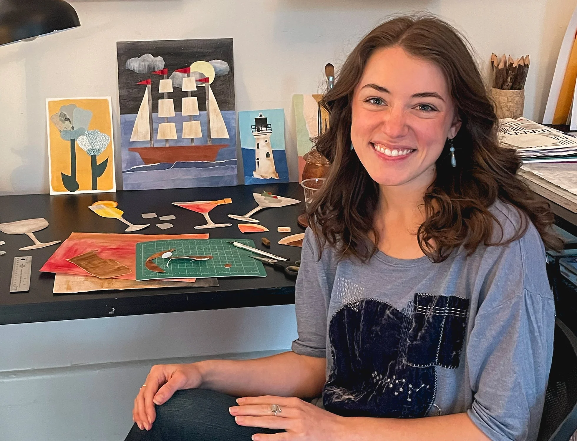 A woman with wavy brown hair, smiling, sitting in front of a table with art supplies and handmade paper cutouts of cocktails. Behind her, there are four small paintings, including a ship sailing at night, a lighthouse, a cactus with flowers, and an abstract. To her right, there are stacks of papers and a container with colored pencils.