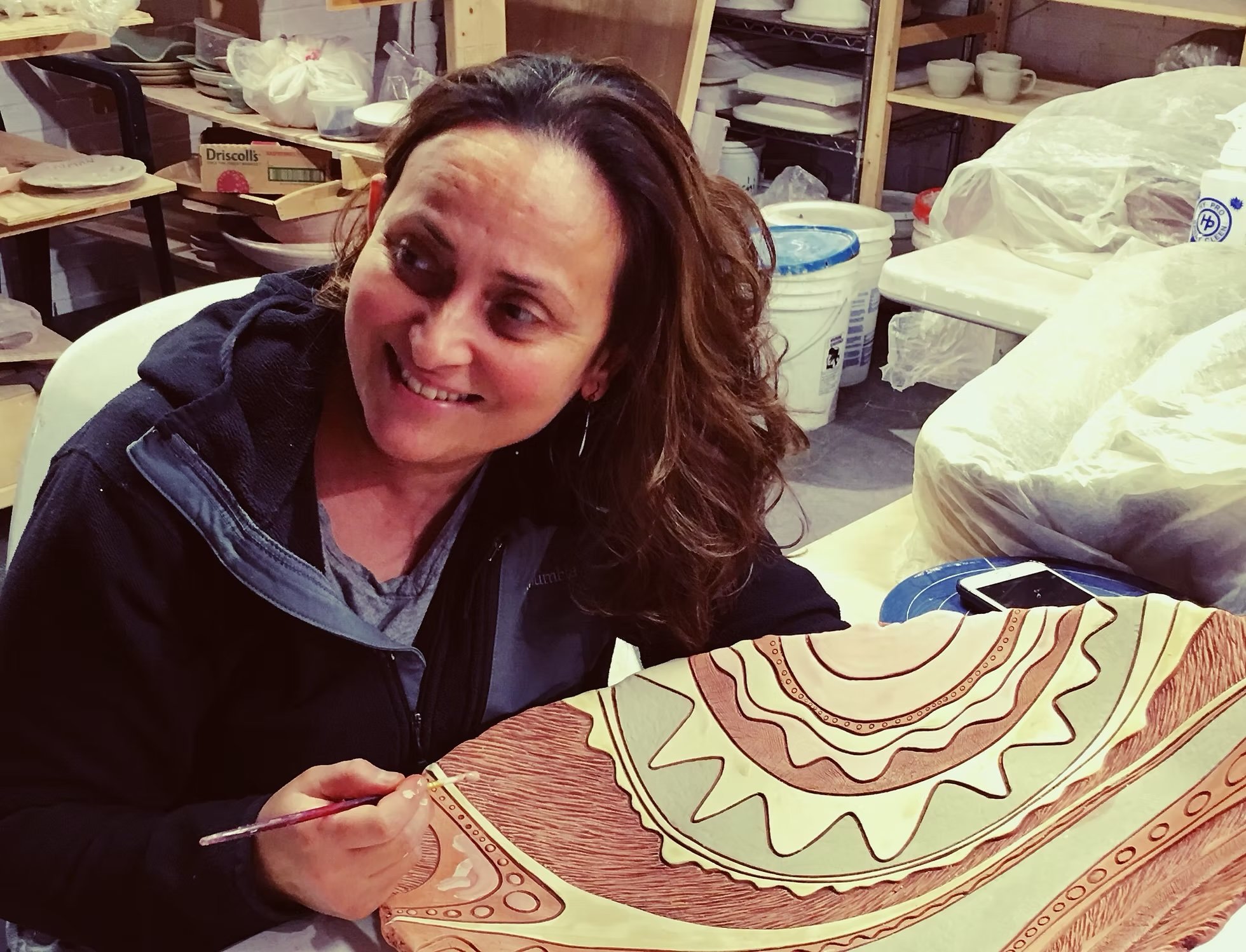 A woman with brown hair, smiling, sitting at a table with an art piece that appears to be a painting or drawing of a pie with a decorative border and design. She is in a workshop or studio with shelves holding pottery and art supplies in the background.