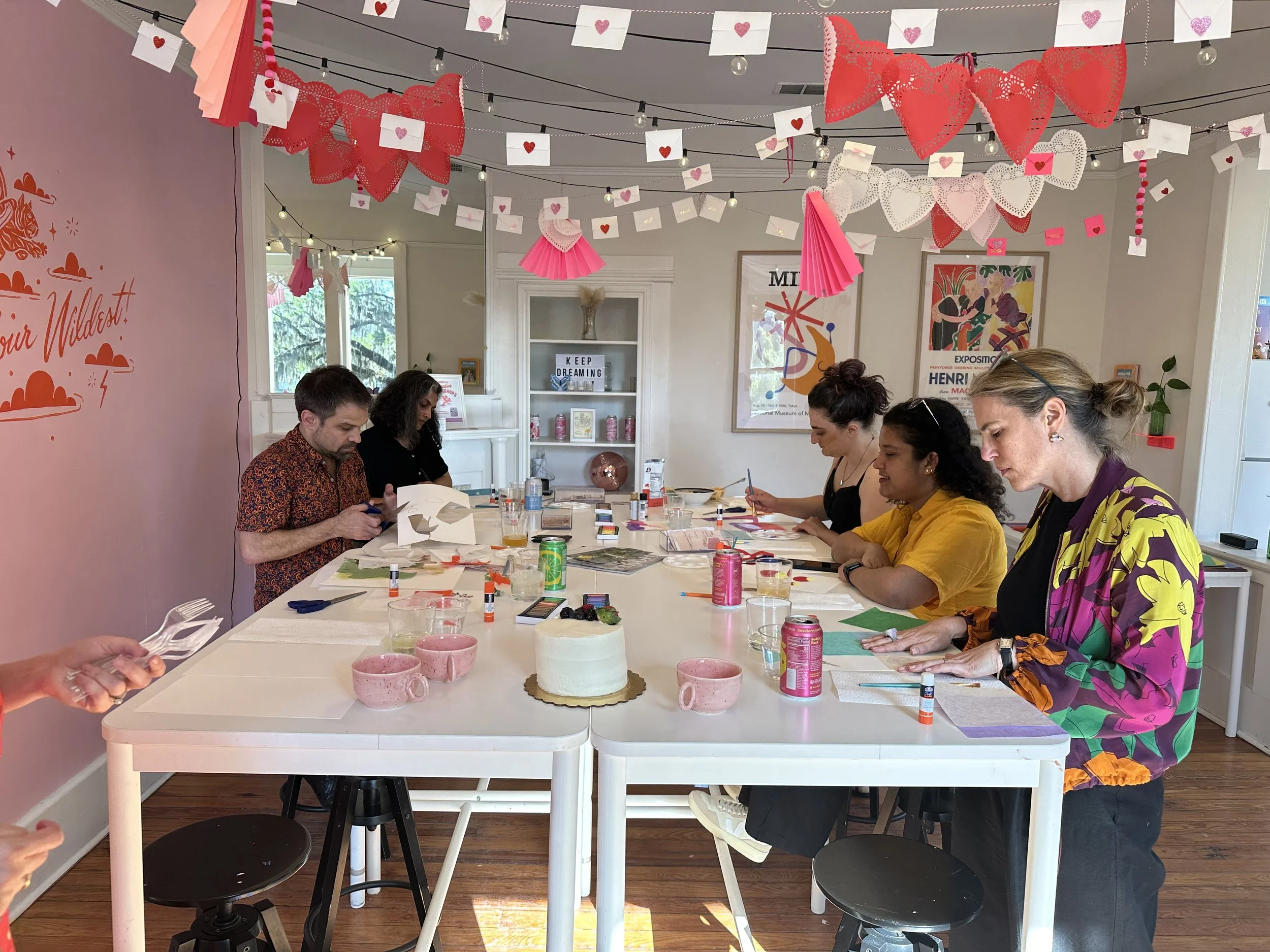 People sitting around a table with craft supplies, a white cake with pink decorations, and pink cups, in a decorated room with pink, white, and red paper decorations hanging from the ceiling.