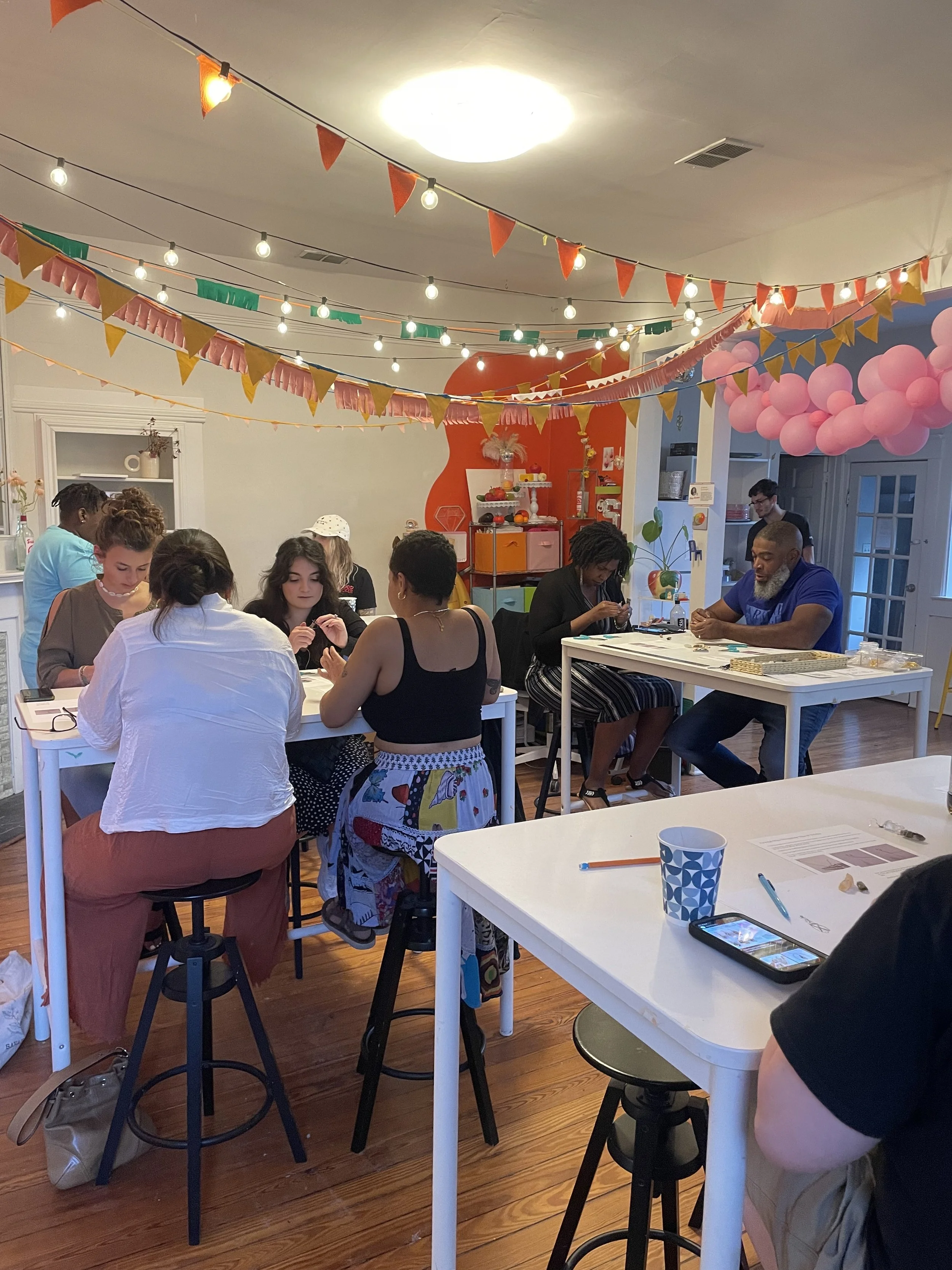 A group of people gathered around tables inside a decorated room with colorful flags and pink balloon decorations hanging from the ceiling. Some are engaged in activity while others are sitting and talking.