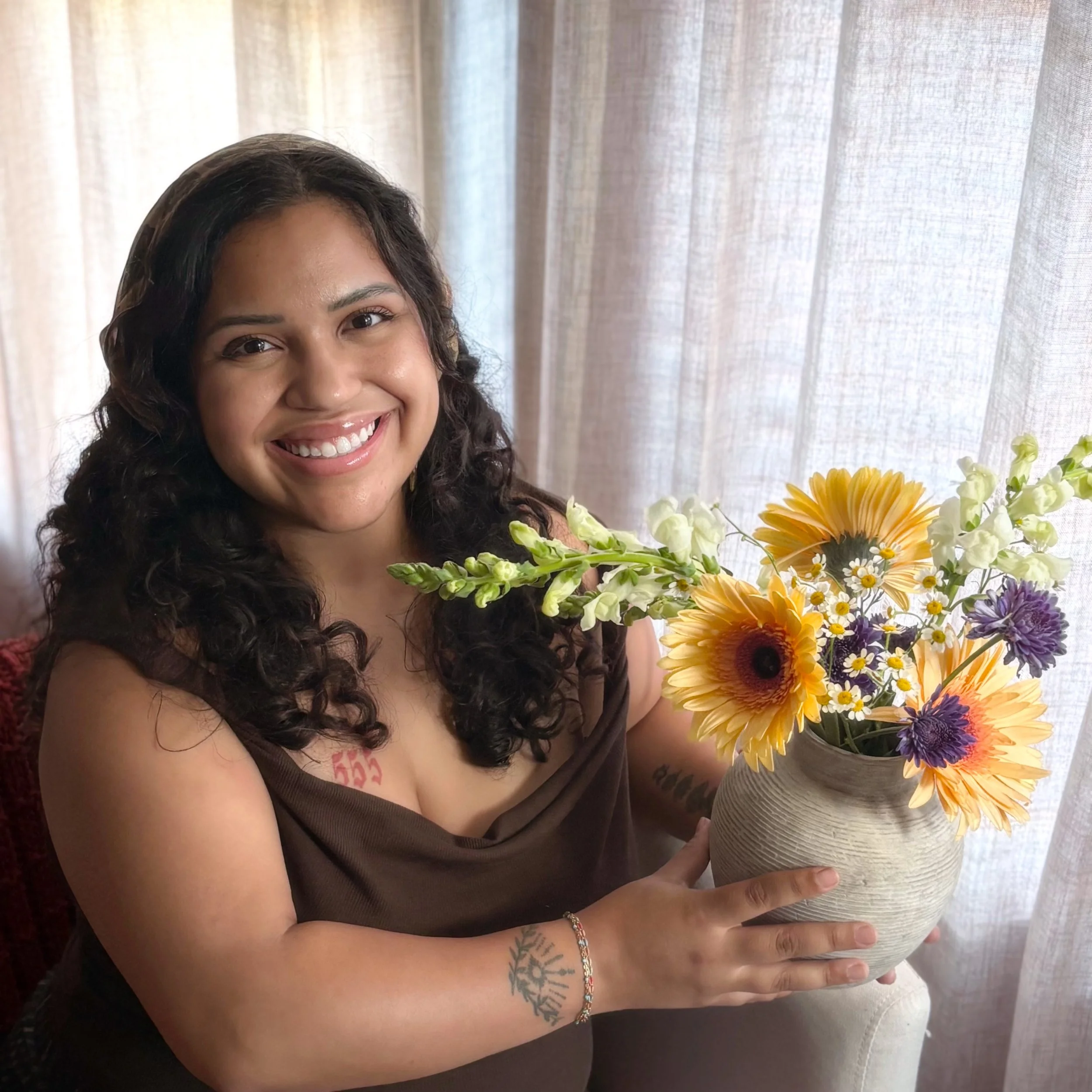Close-up of a smiling woman with curly dark hair holding a large bouquet of colorful flowers in front of a sheer curtain.