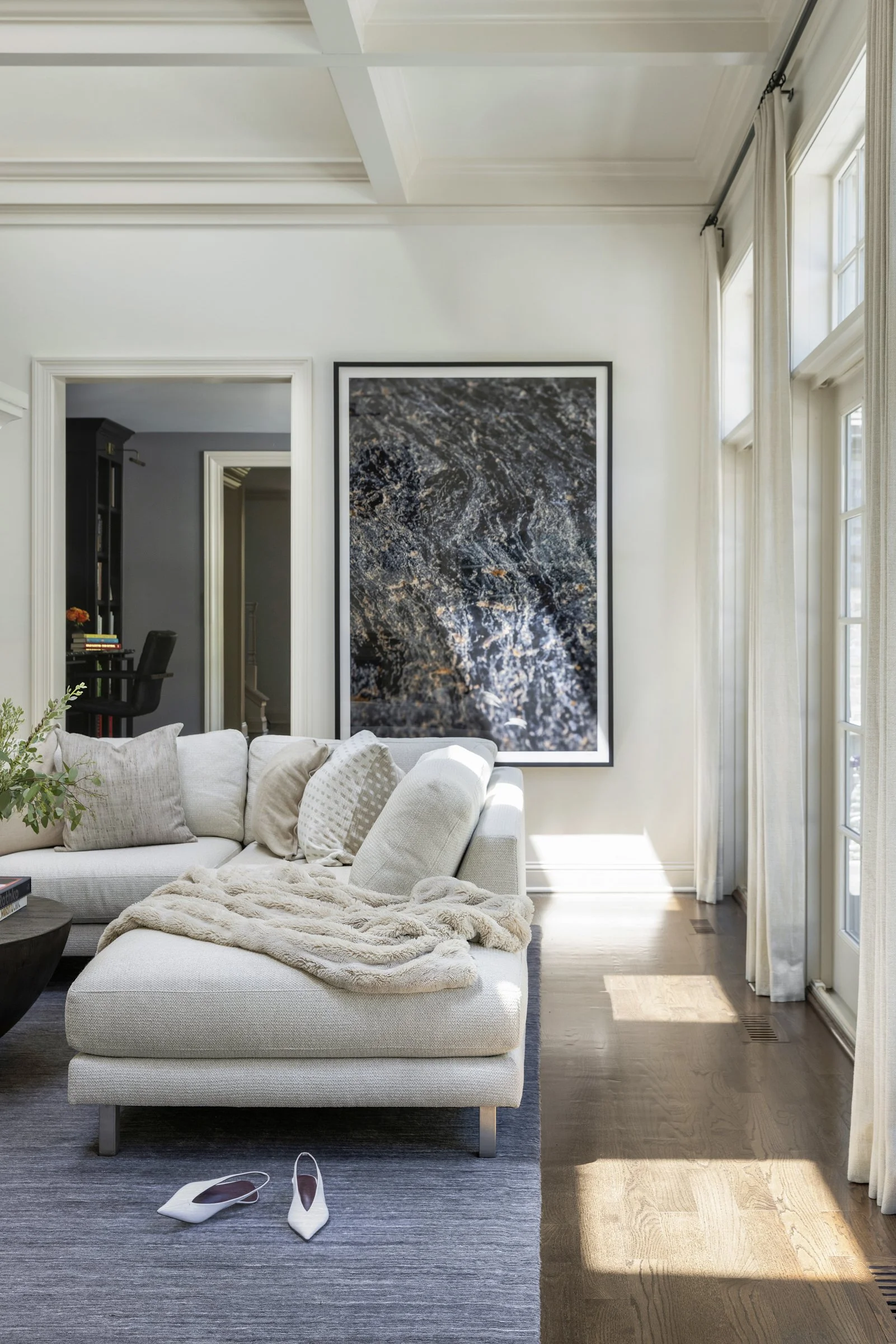 Modern, luxury living room with white sofa and throw blanket, beige pillows, closed-toe heels on a grey rug, large window with curtains, framed artwork on the wall, hardwood floor, and a doorway leading to a darker room.