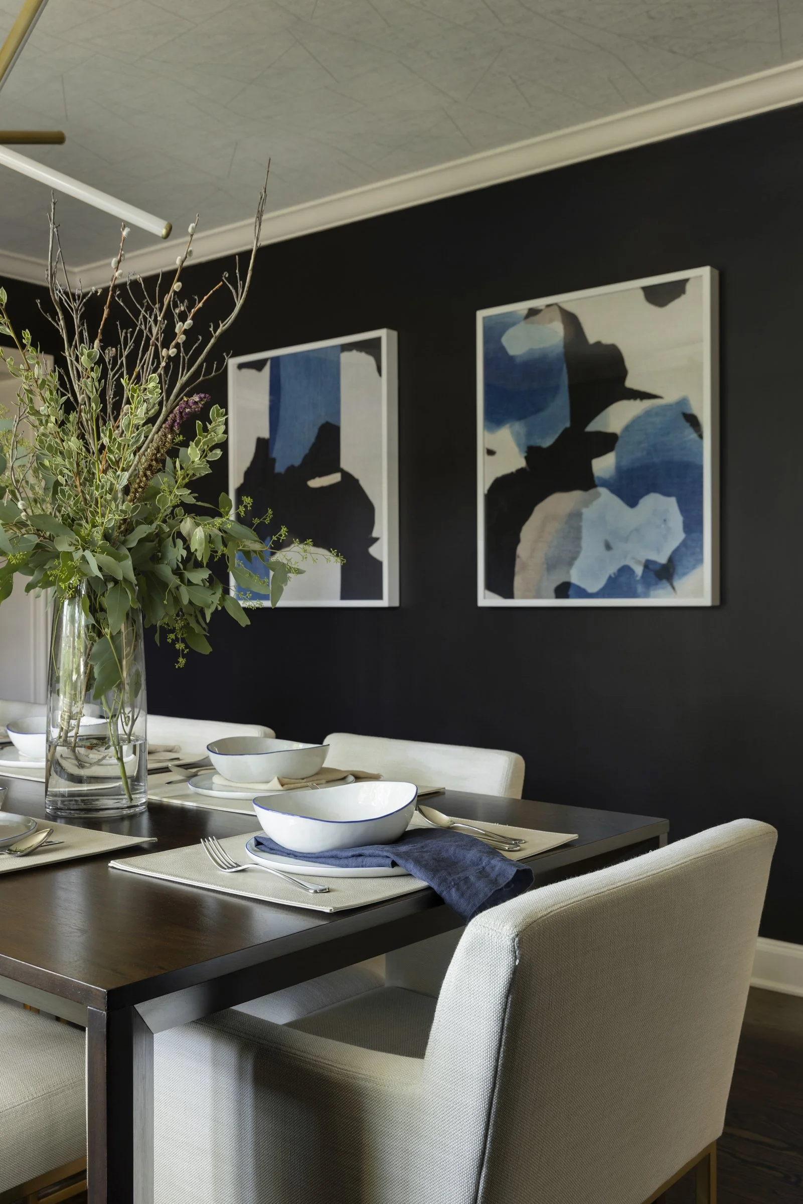 A modern dining room with a dark wood table set with white bowls, silverware, and napkins, decorated with a large floral arrangement in a glass vase, with abstract artwork on a dark wall in the background. Beautiful luxury interior design.