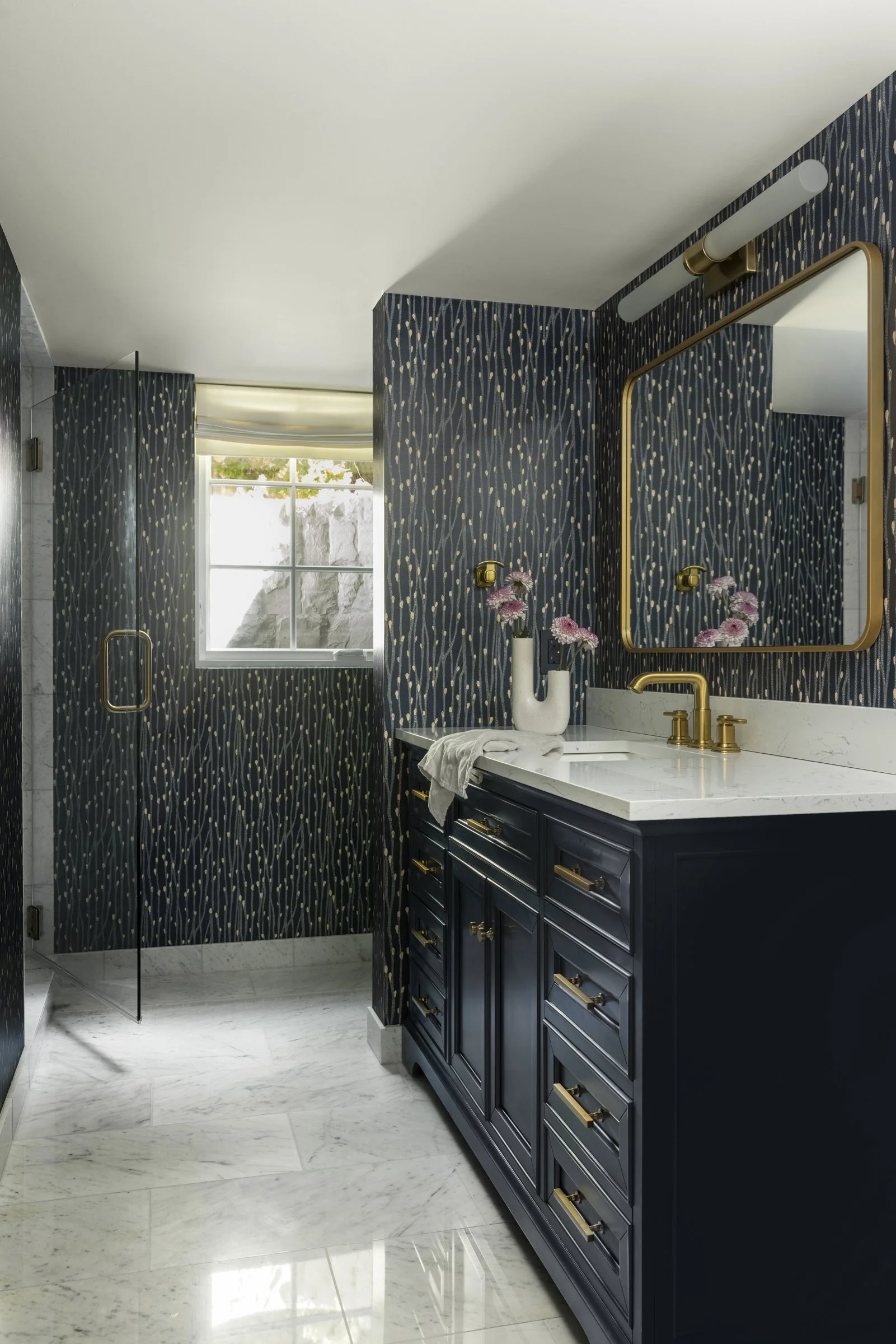 A bathroom with a navy blue vanity and gold hardware, a white marble countertop, a vase with pink flowers, a large mirror with a gold frame, a modern wall-mounted light fixture, patterned navy wallpaper, a window allowing natural light, a glass showe