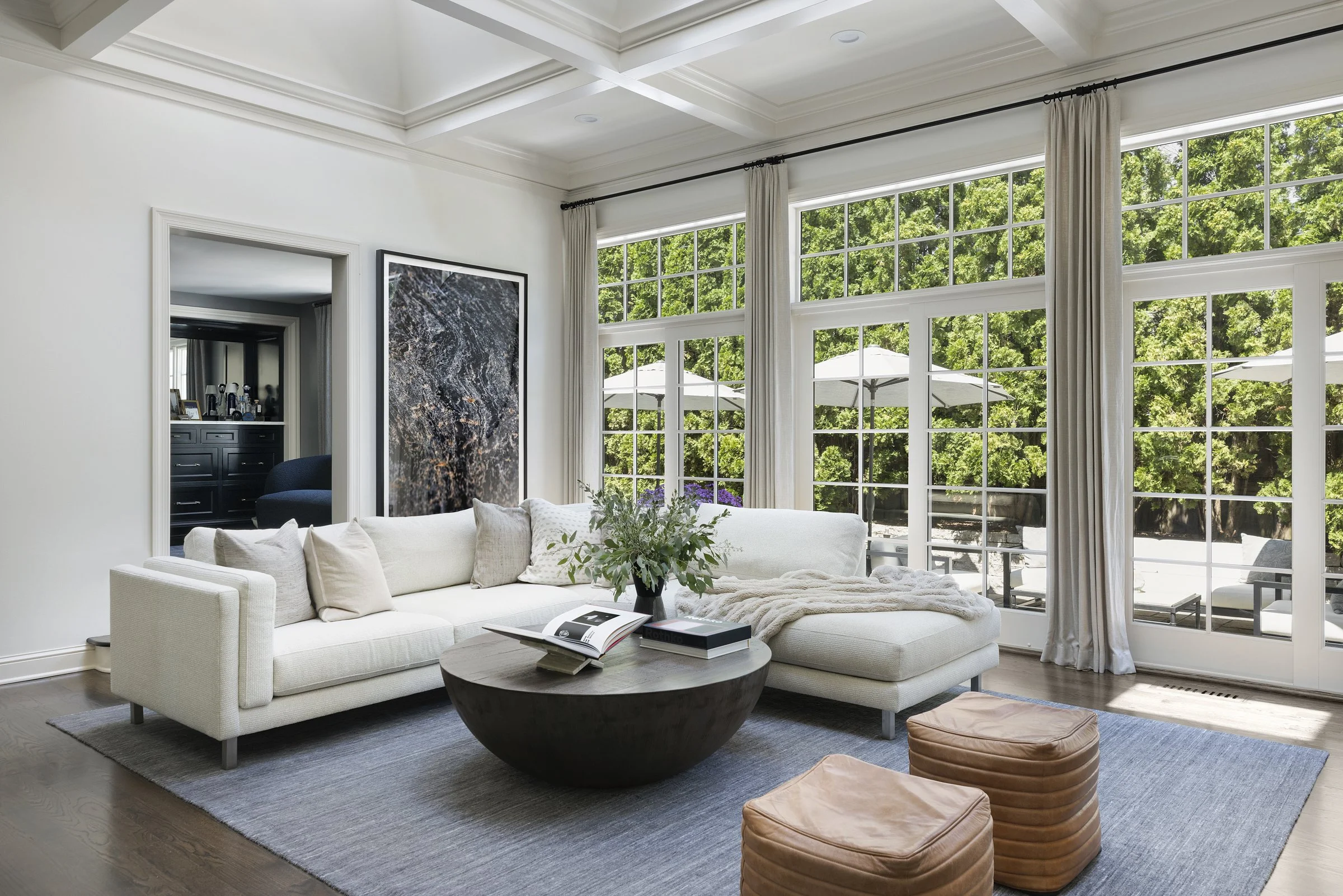 Bright modern, luxury living room with large window walls showing outdoor patio umbrellas and trees, white sofa, round coffee table with books and plant, beige ottomans, gray rug, and doorway to darker-colored room.