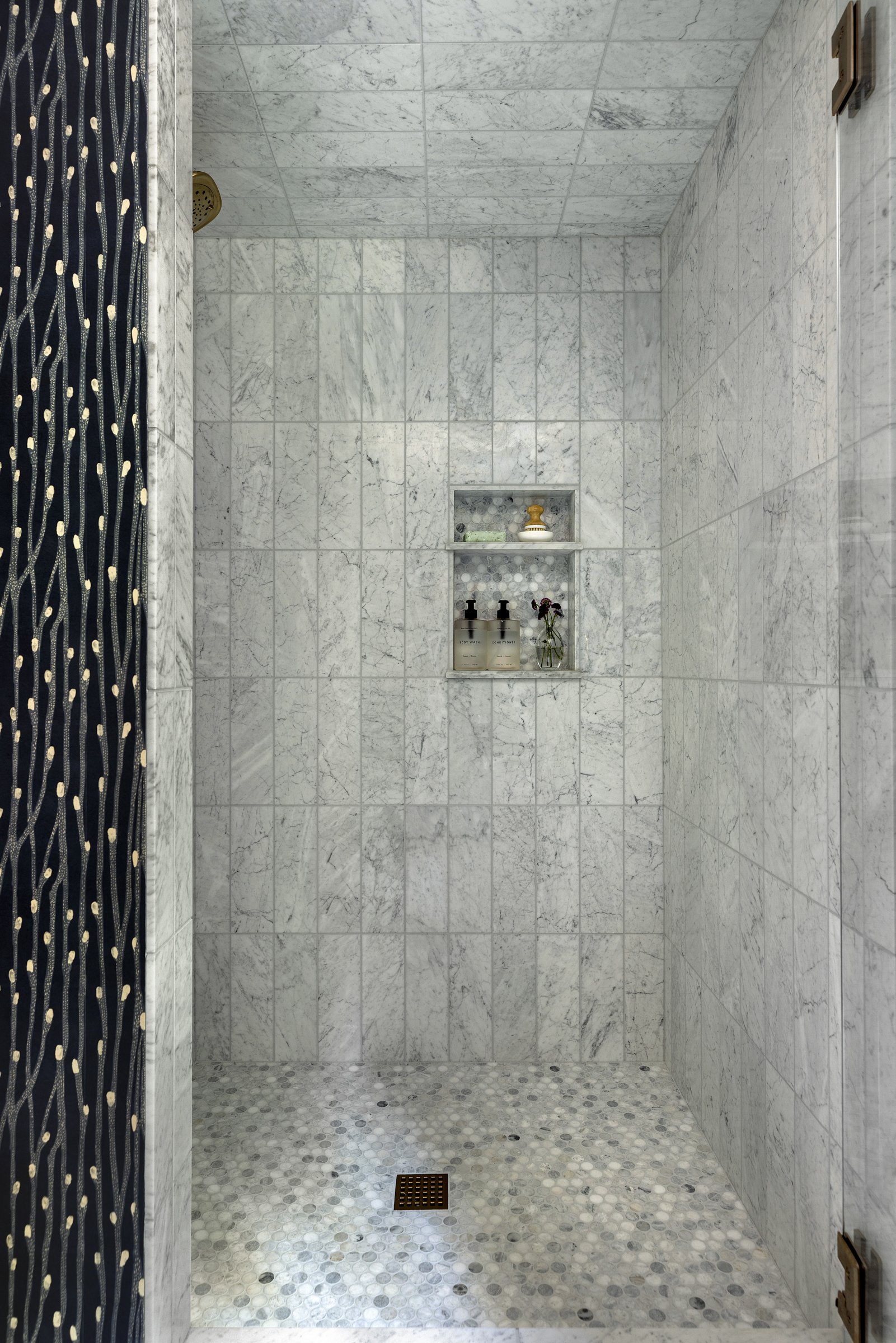 Shower area with white marble tile, a built-in shelf with toiletries, a drain on the floor, and a dark shower curtain with a pattern. Beautiful bathroom renovation.