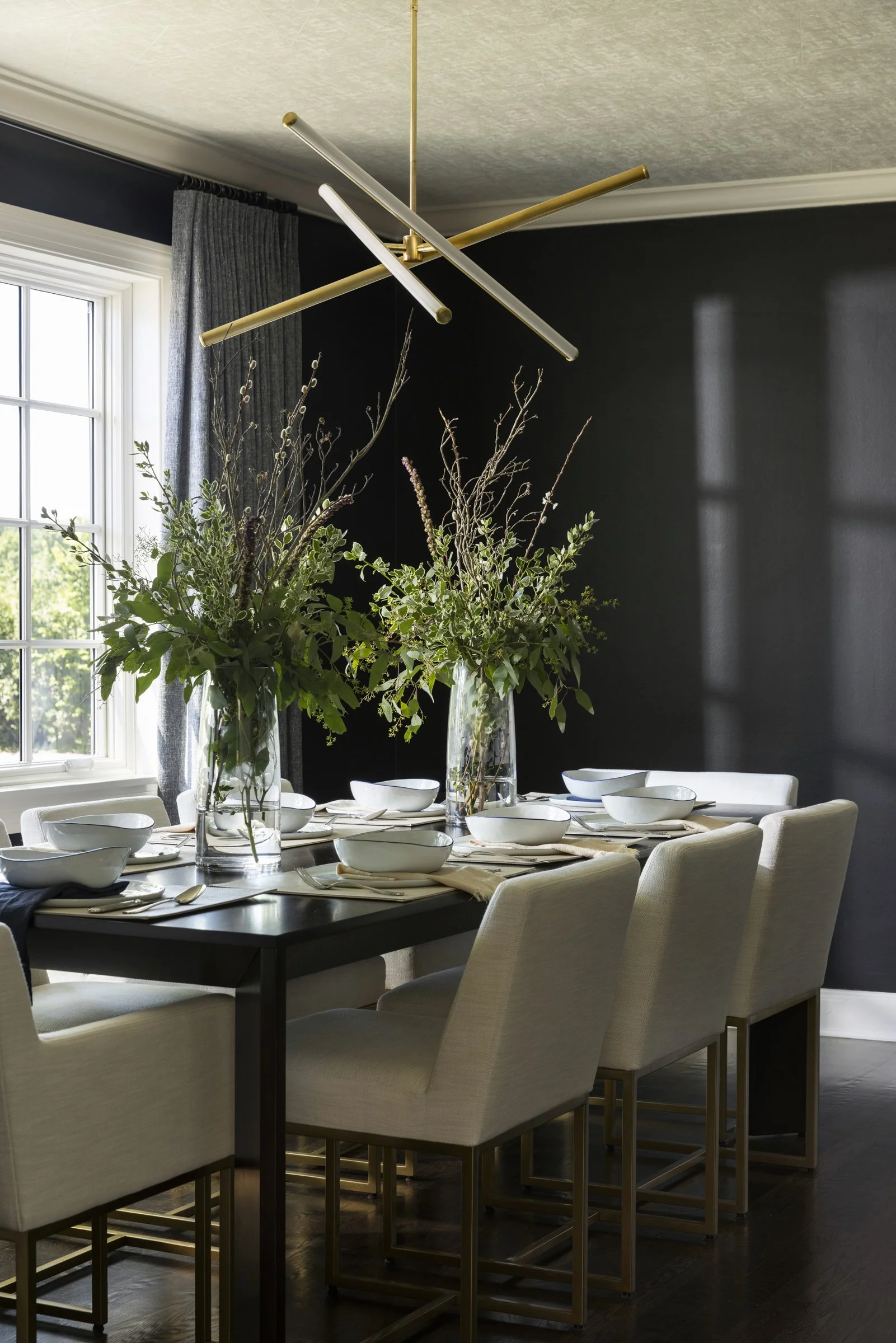 A dining room with a black table set with white bowls and napkins, surrounded by beige upholstered chairs, and decorated with two large glass vases holding leafy green branches; a window with gray curtains is visible, and a modern ceiling light fixtu