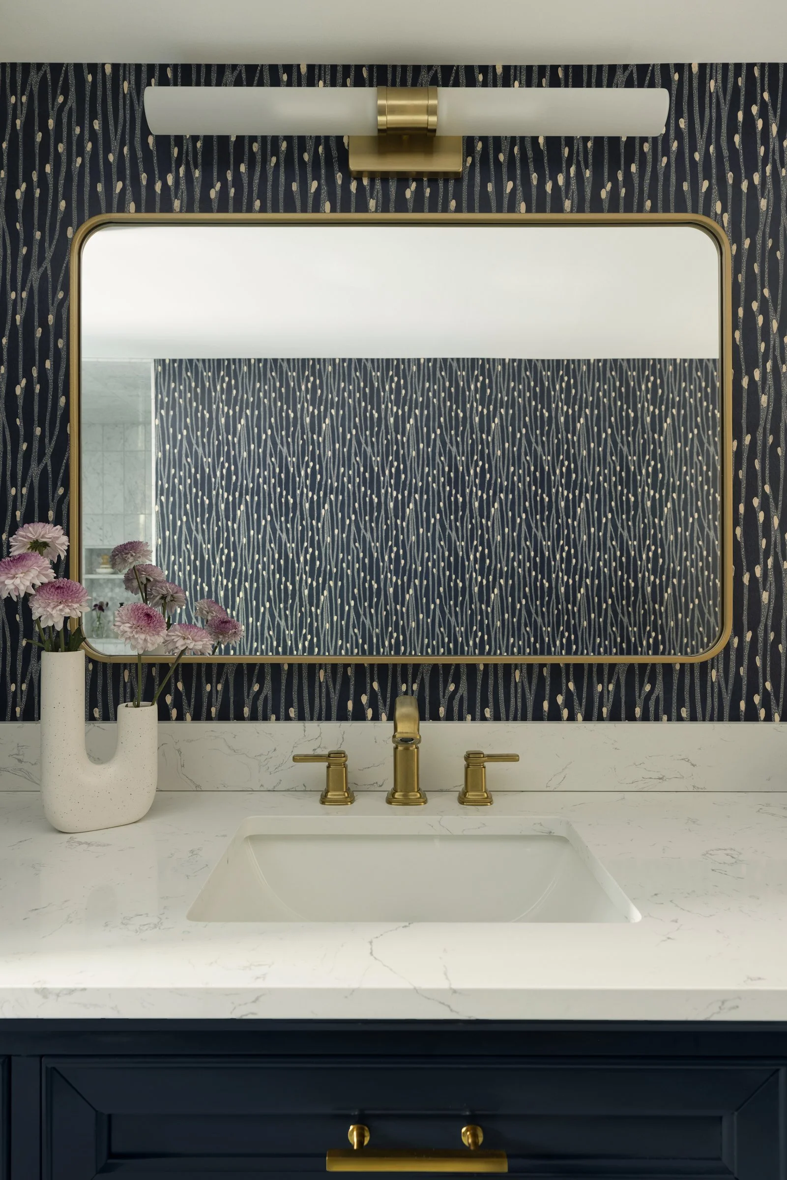 A blue luxury bathroom vanity with a white marble countertop, gold fixtures, a rectangular undermount sink, a navy blue cabinet with gold knobs, a large wall mirror with gold trim, a modern wall light fixture, and a white vase with pink flowers.