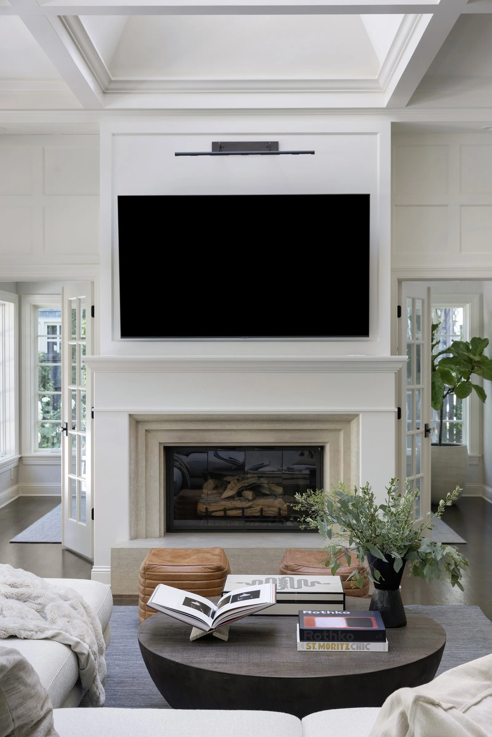 Living room with a fireplace, large flat-screen TV, framed by white molding. French doors flanking the TV open to an outdoor space, with a plant visible on the right. A wooden coffee table holds books and an open photo album, with a gray rug undernea