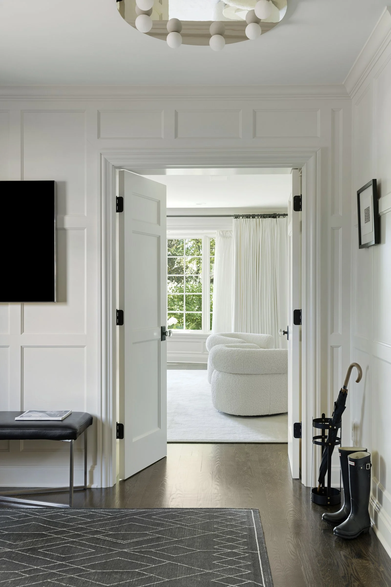 Entryway view leading into a living room with a window, white curtains, seating, and a ceiling light fixture with round bulbs. A pair of black rubber boots and umbrella are by the wall.