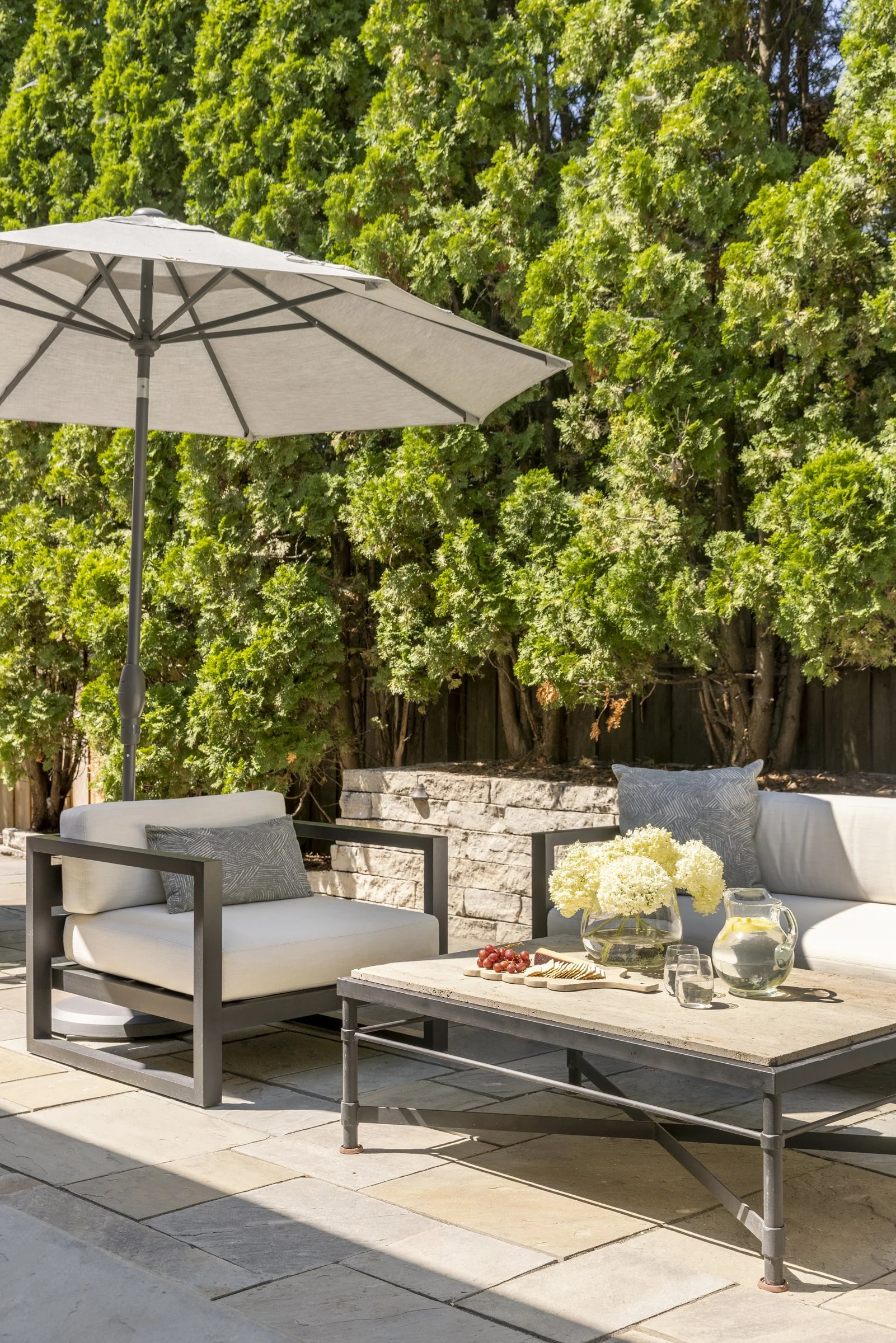 Outdoor patio seating area with white cushioned chairs, a stone firepit, a coffee table with a vase of white flowers, grapes, cheese, and wine glasses, shaded by a large grey umbrella, with a green leafy hedge in the background.