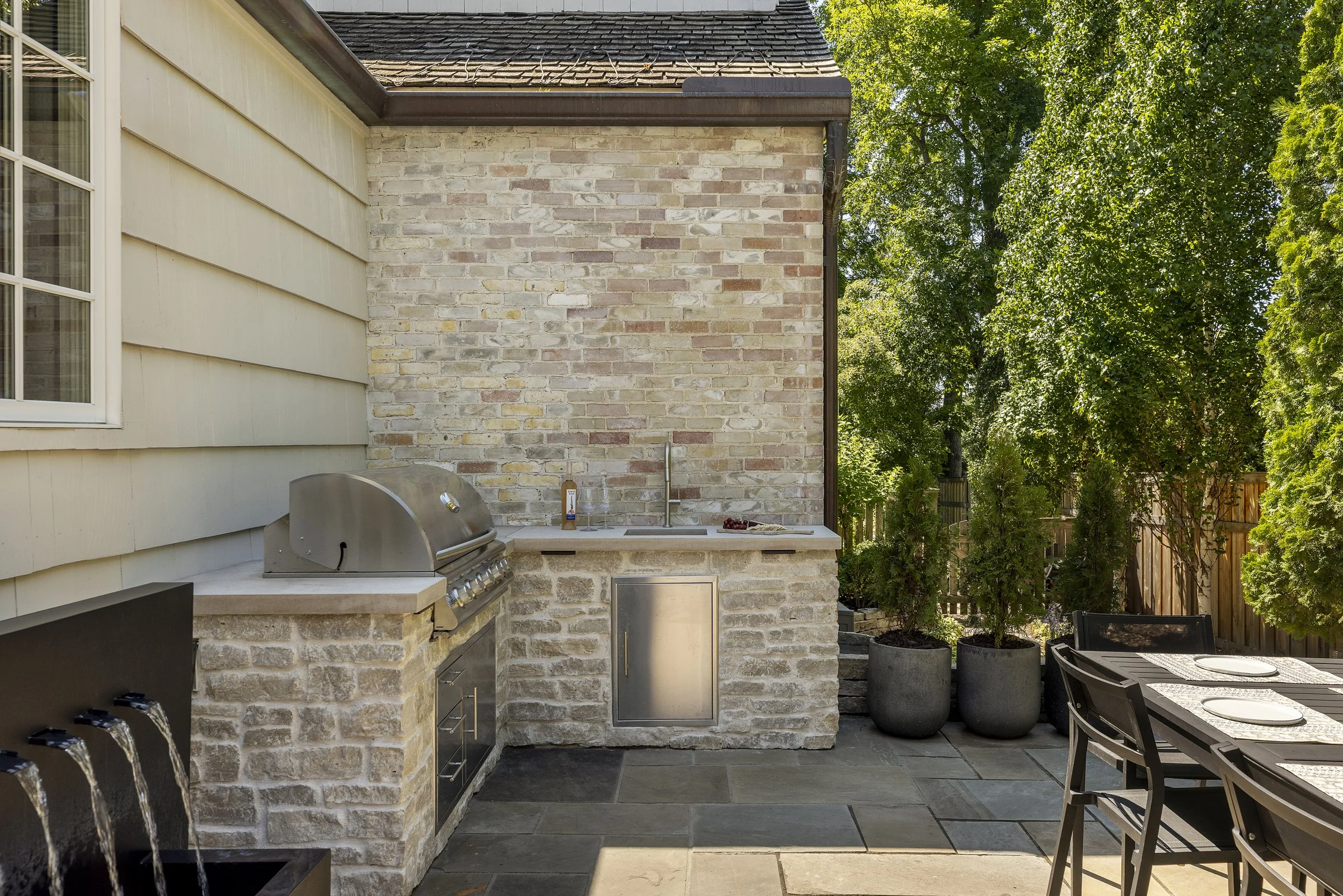 An outdoor patio area with a built-in grill, a kitchen countertop, a sink, and potted plants with trees in the background.