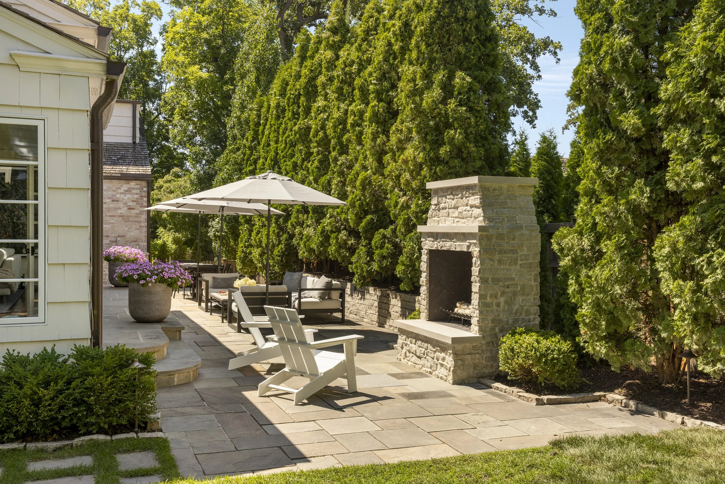 A backyard patio with outdoor furniture, including white chairs, a large umbrella, a stone fireplace, and potted pink flowers, surrounded by tall green trees and landscaped bushes.