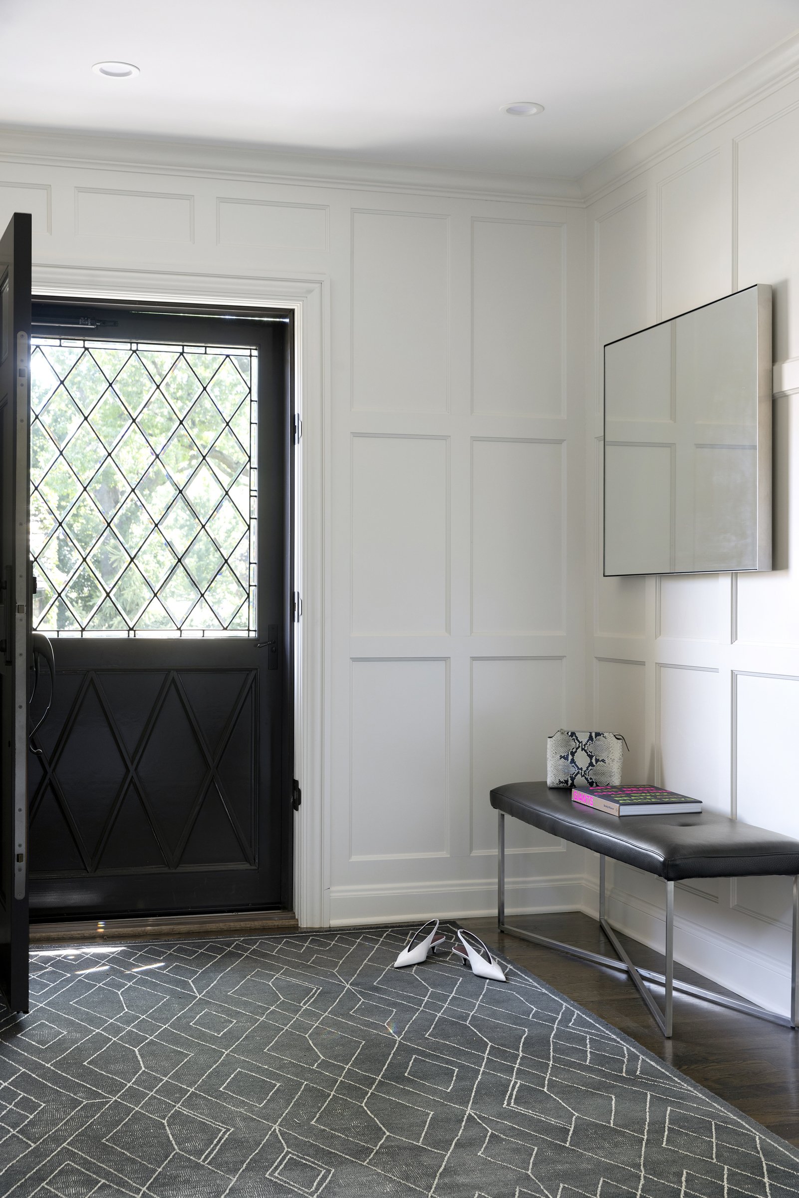 Interior entryway with a black metal door featuring a diamond-patterned window, white paneled walls, a gray bench with black leather cushion, a small white-patterned decorative item, a book, and a pair of white high-heeled shoes on a geometric gray r