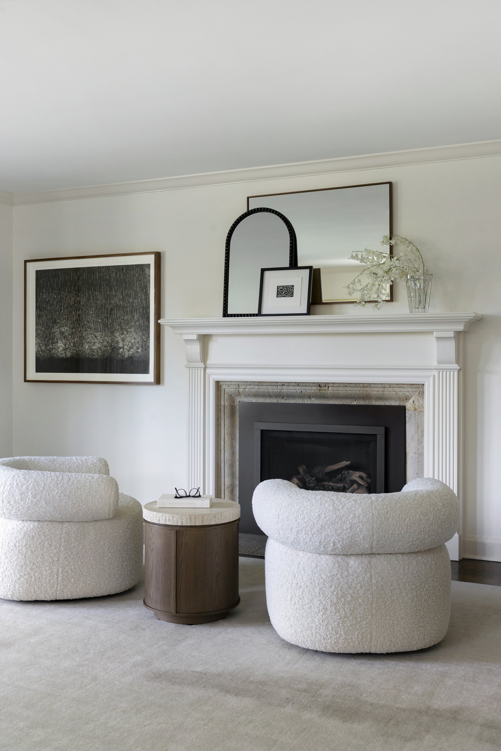 Living room with two white textured armchairs around a small round wooden side table, a fireplace with white mantel, framed pictures, mirror, and a glass vase with white flowers on the mantel, beige carpet, and abstract artwork on the wall.