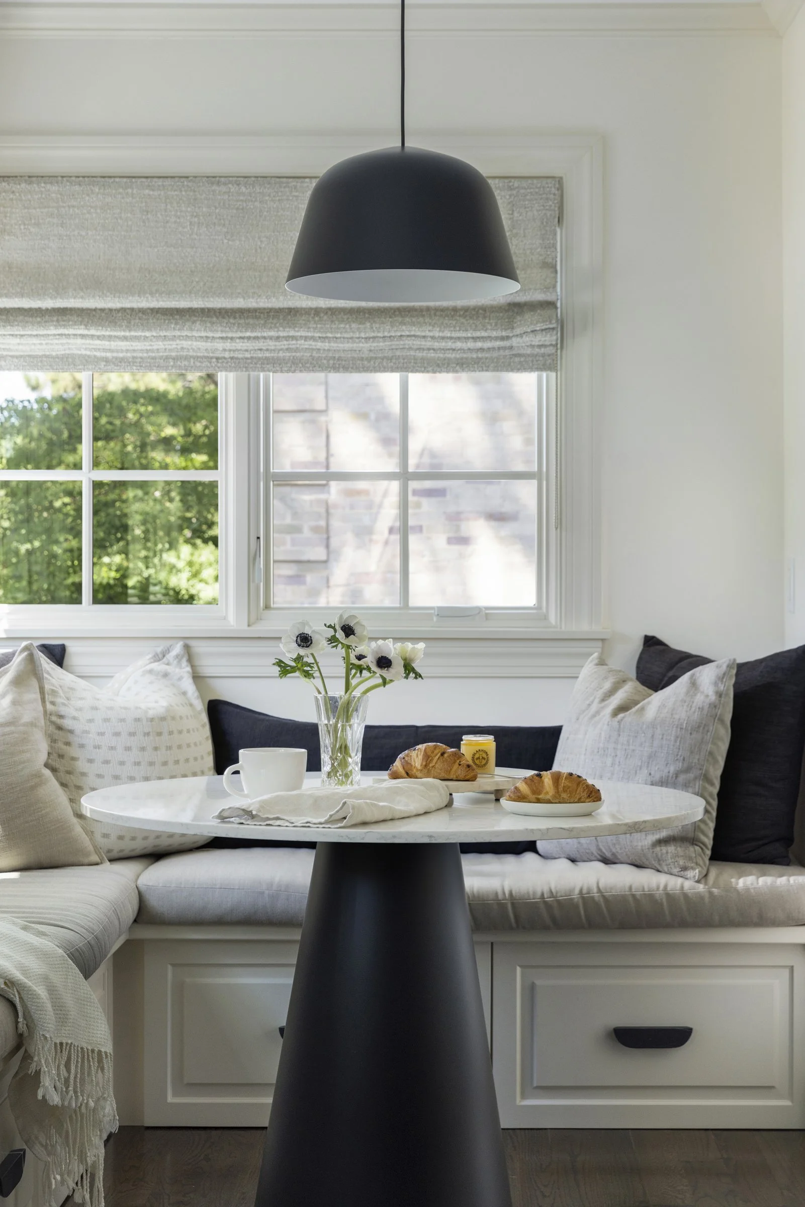 Bright luxury kitchen nook with a window, a black pendant light, and a small round table with breakfast items, including croissants, a white mug, a jar of honey, and a vase with white flowers, surrounded by throw pillows on the sofa.
