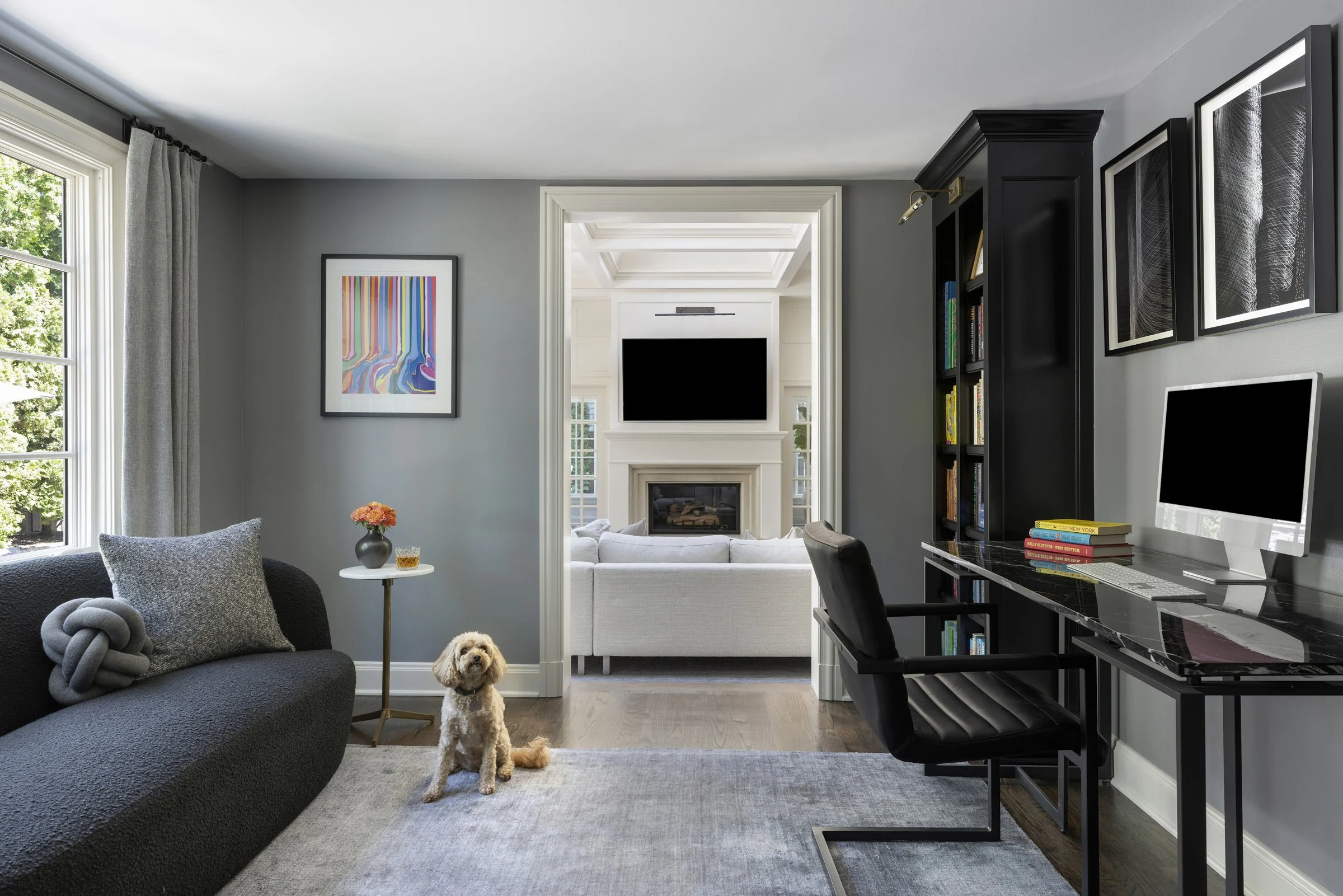 Home office with large window, gray walls, abstract art, black bookshelf, computer, and dog sitting on rug.
