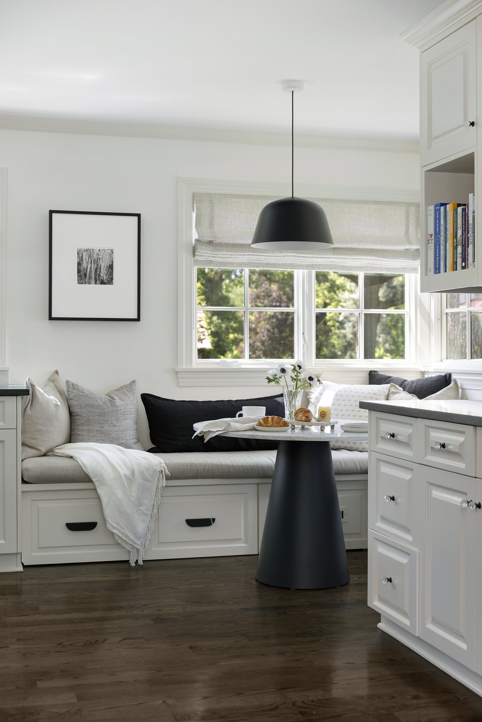 A bright, modern kitchen and living area with white cabinets, hardwood floors, a black pendant light, a window with Roman shade, and a white daybed with gray and black pillows, a black side table, and a round table with breakfast items.