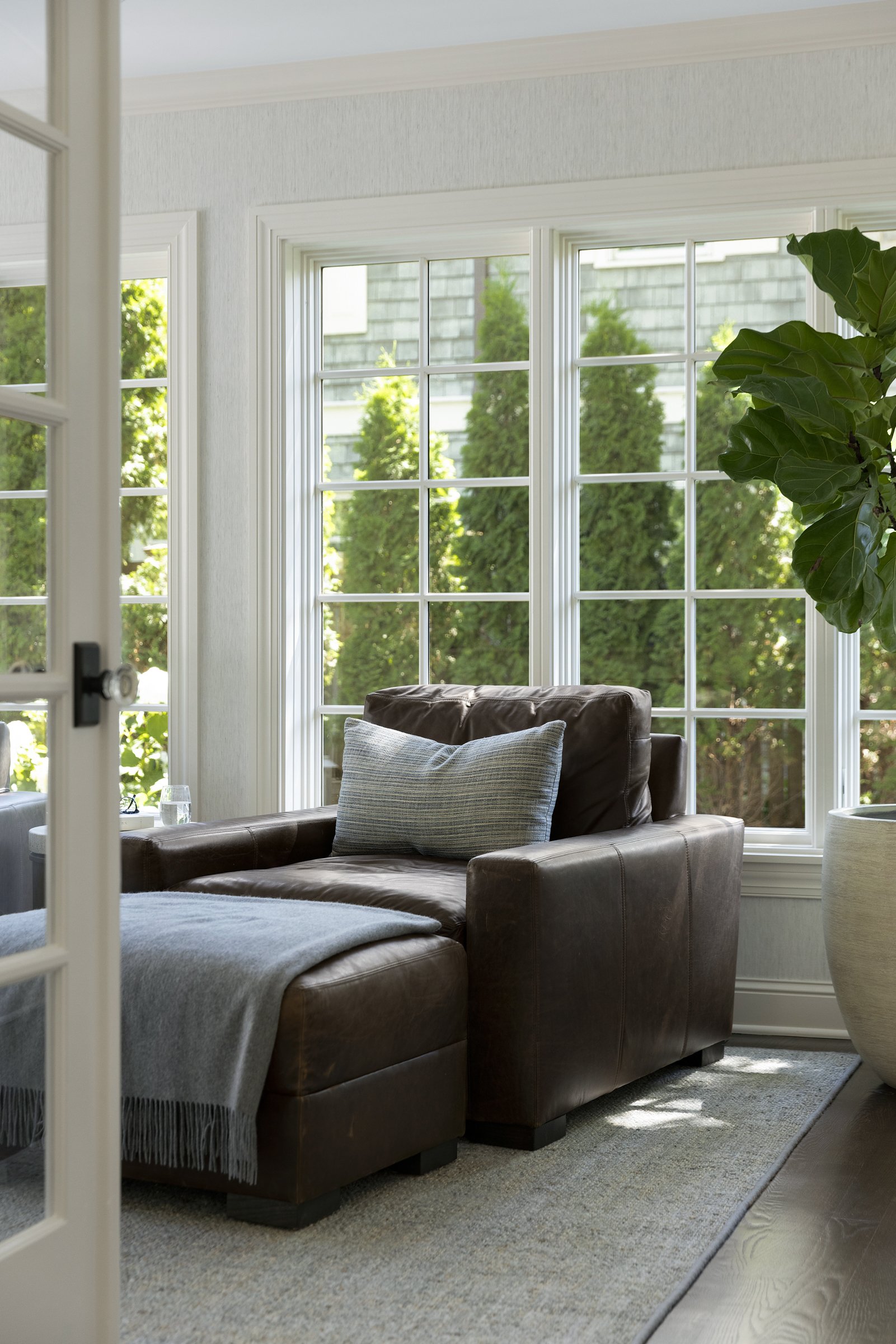 Sunroom and living room with large windows, a leather sofa with a throw blanket, a decorative pillow, a large potted plant, and natural light coming in from outside. Beautiful luxury interior design.