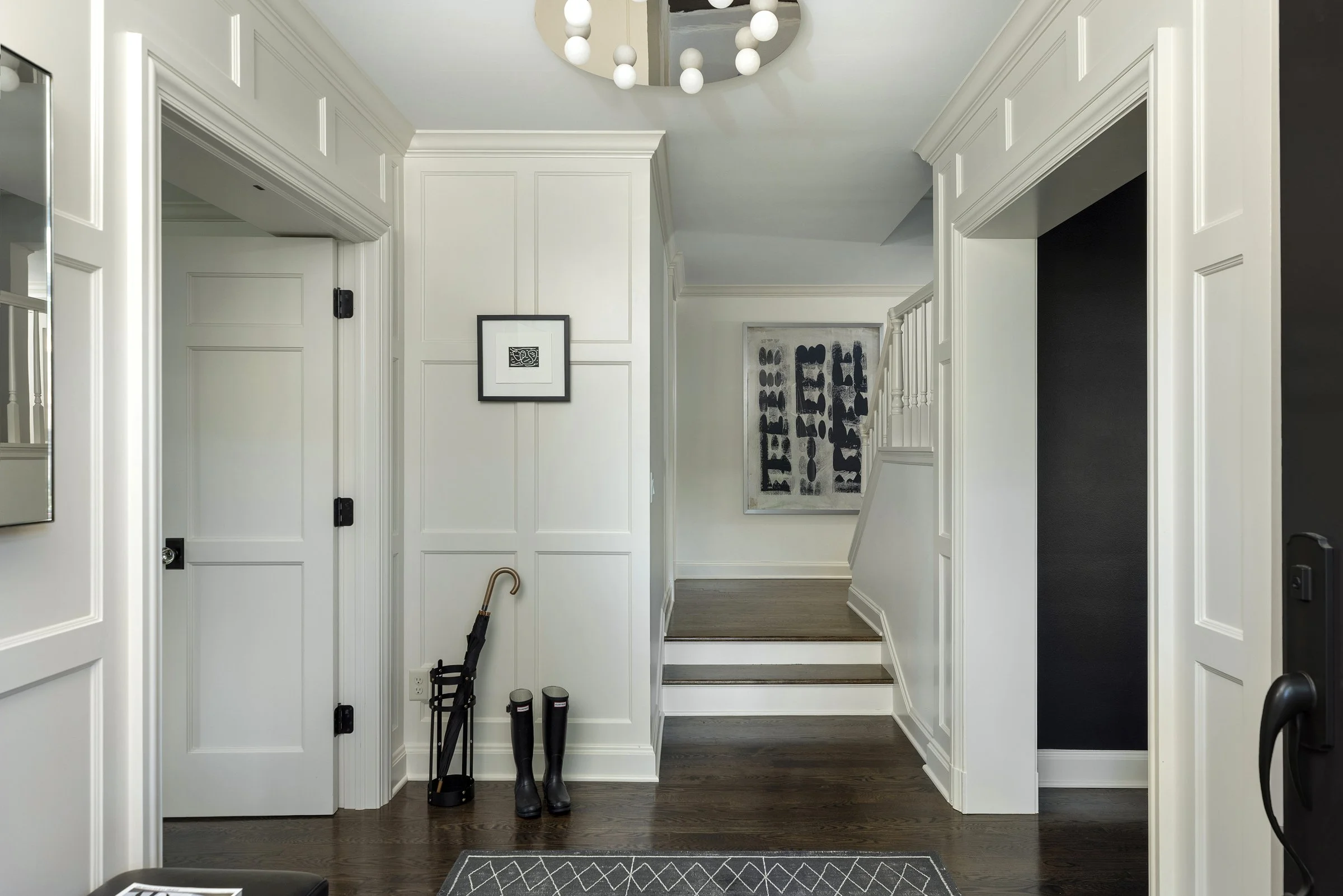 Entryway with white paneled walls and dark wood flooring, featuring a black umbrella stand with umbrellas, a framed black and white artwork, and a staircase with white banister leading upward. There is a modern chandelier with spherical bulbs hanging