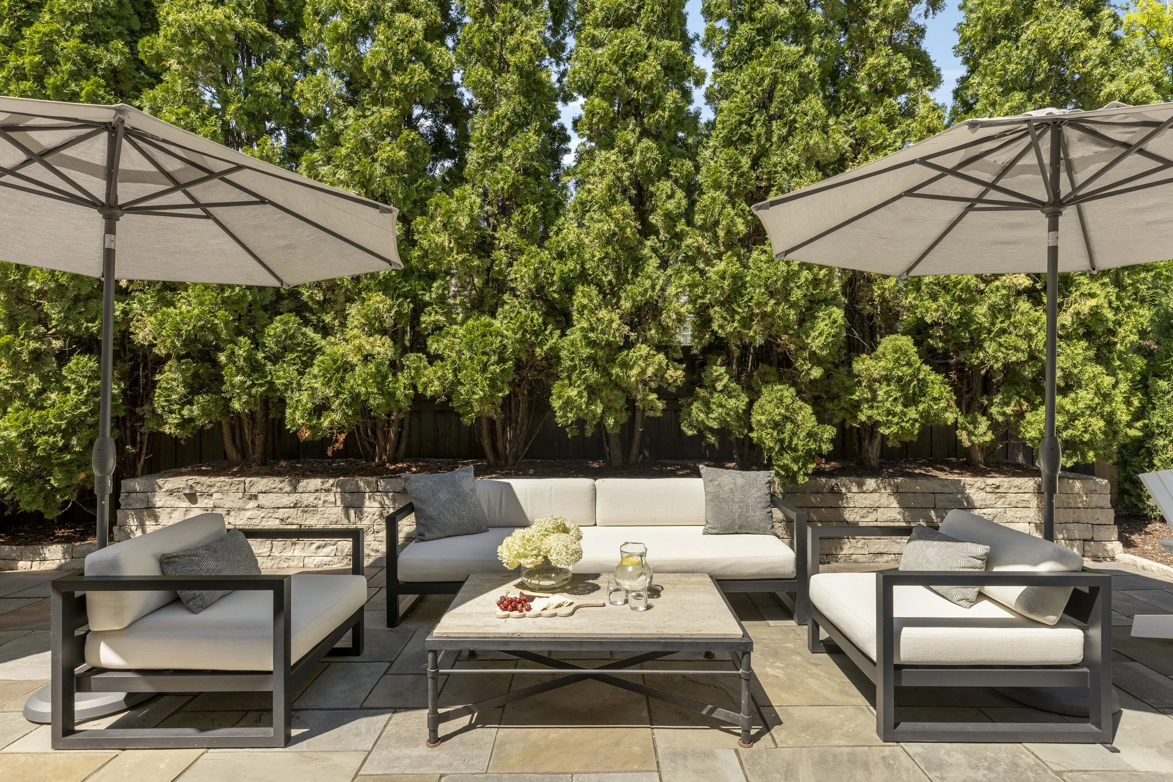 Outdoor patio area with white cushioned seating, a coffee table with floral decoration, two large umbrellas, and a backdrop of tall green trees and a partially visible fence.