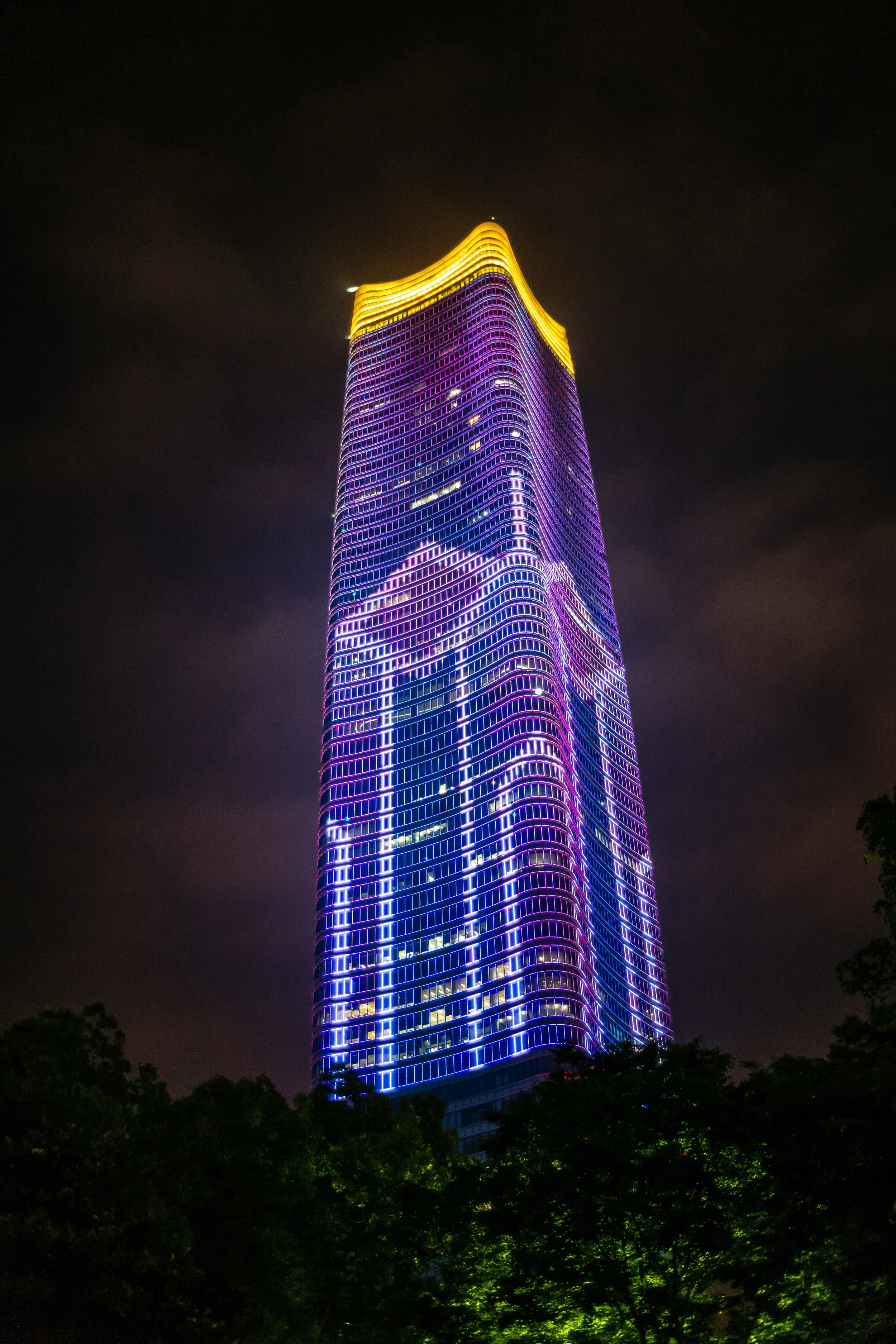 A tall skyscraper at night illuminated with vibrant purple, blue, and yellow neon lights, with dark clouds in the background and green trees at the bottom.