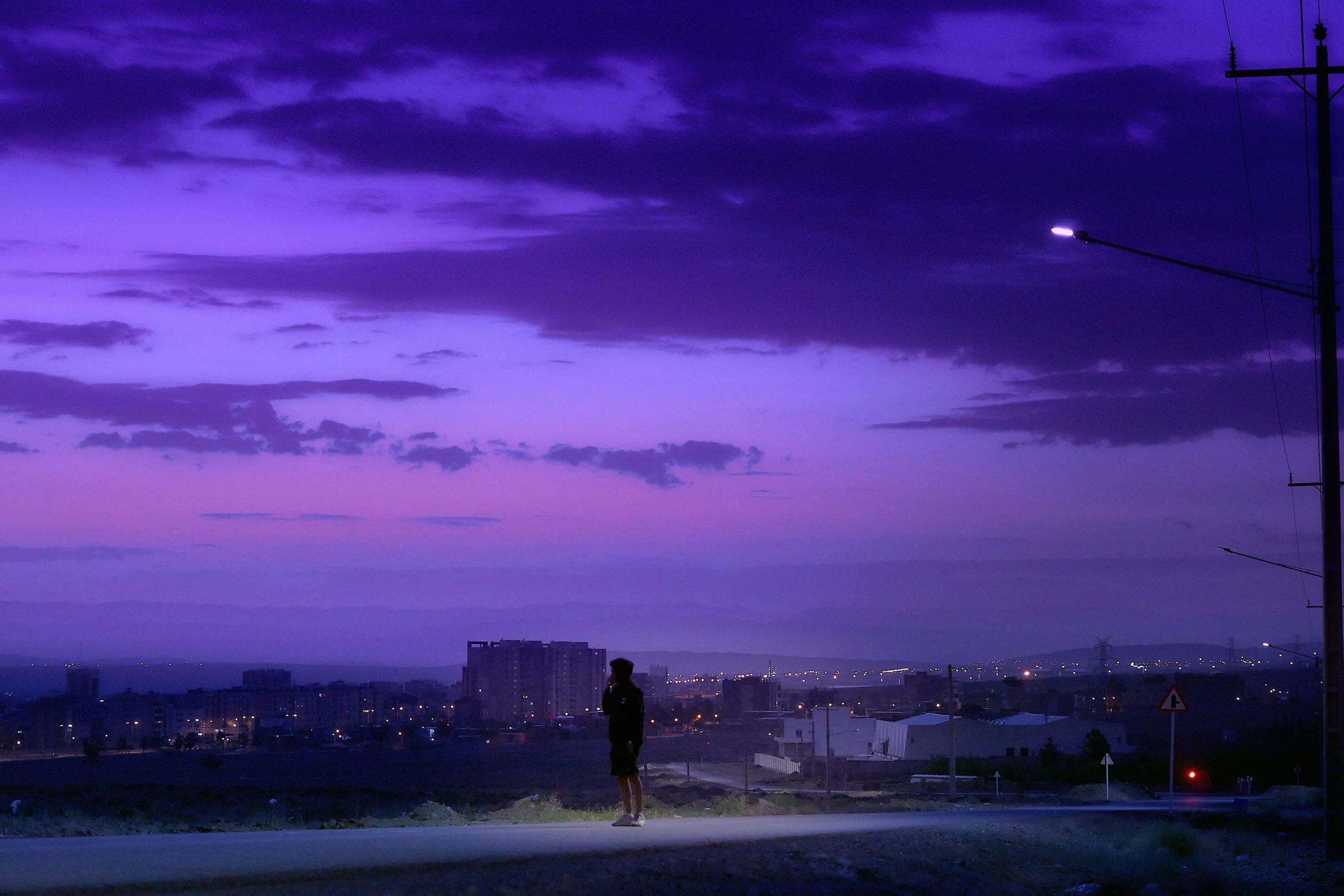 Person standing on the side of the road during twilight with purple and blue clouds in the sky and city lights in the distance.