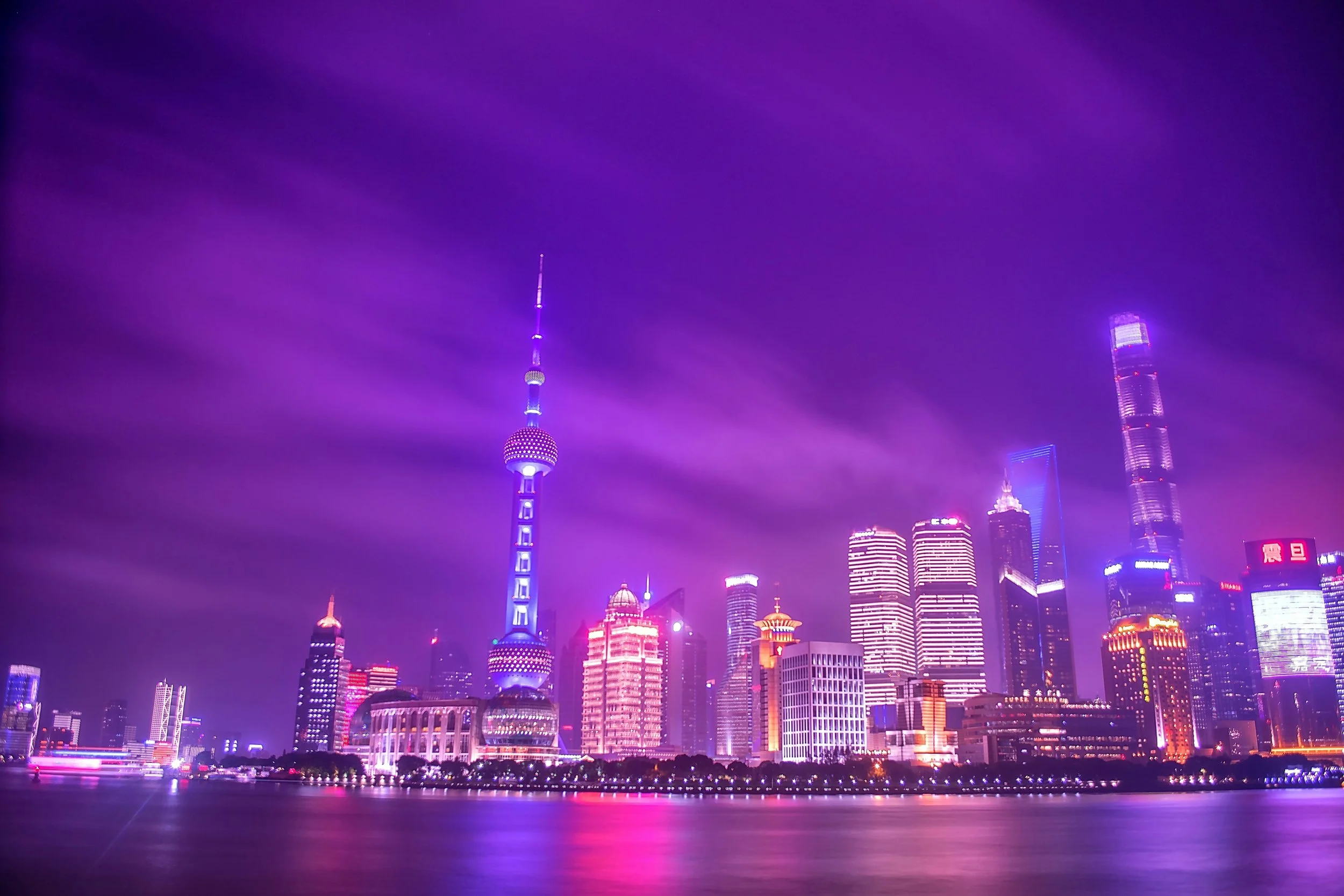 Nighttime city skyline of modern skyscrapers illuminated with colorful purple, pink, and blue lights, reflected on the water below, with a purple foggy sky.