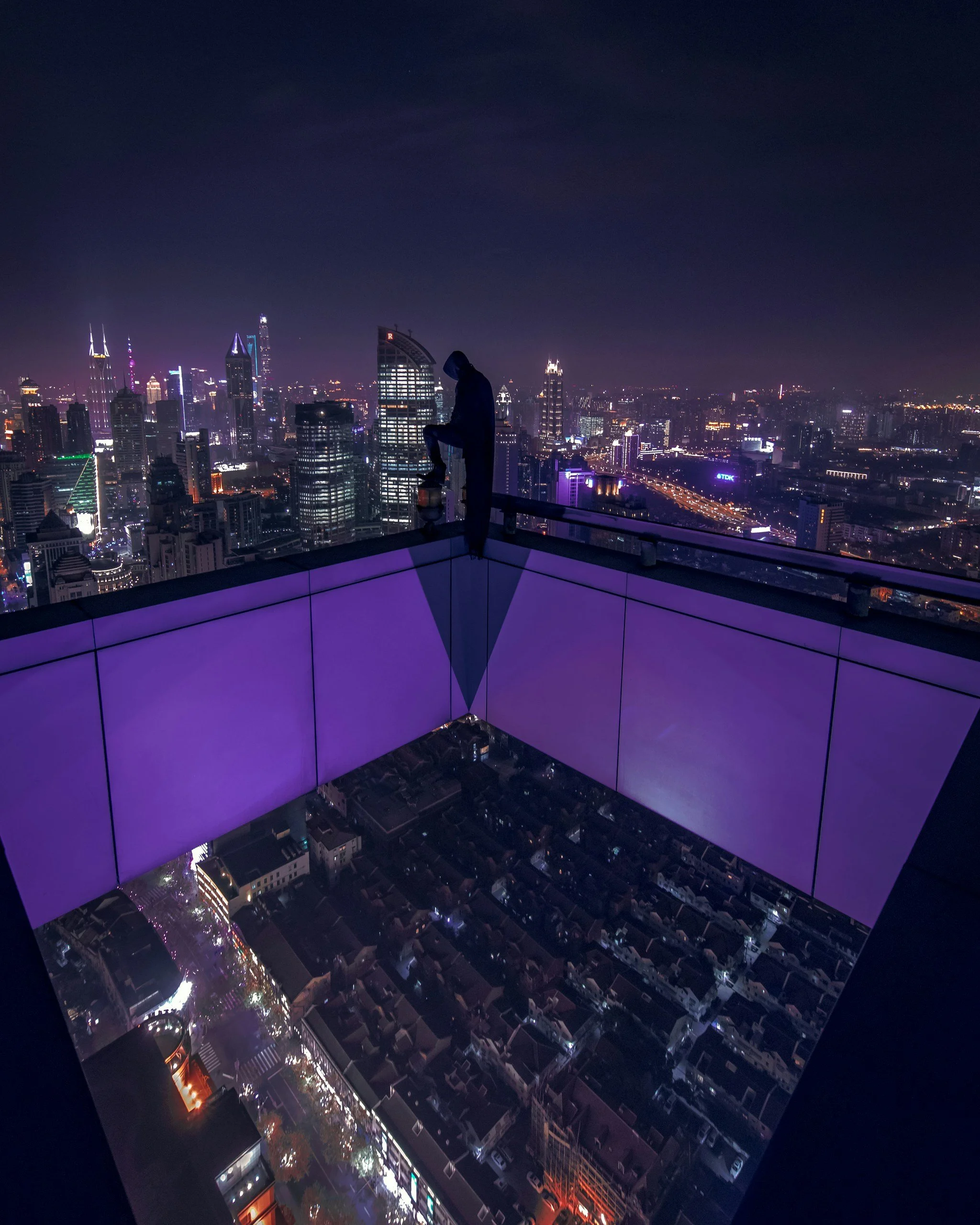 Nighttime cityscape view from a rooftop with a person in a hoodie sitting on the edge, with illuminated buildings and streets in the background.